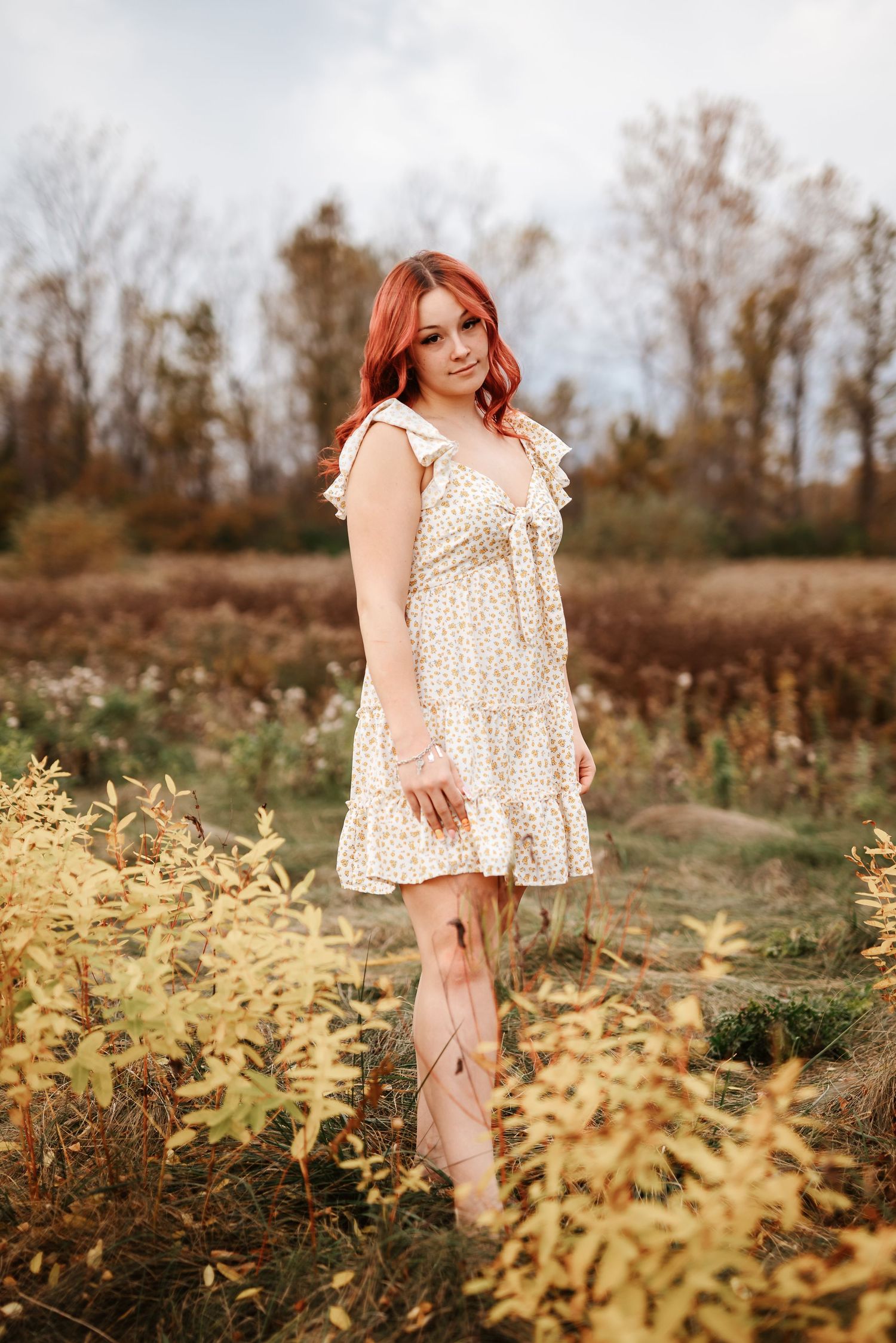 Young woman with red hair in a floral sundress posing in an autumn field with golden wildflowers.