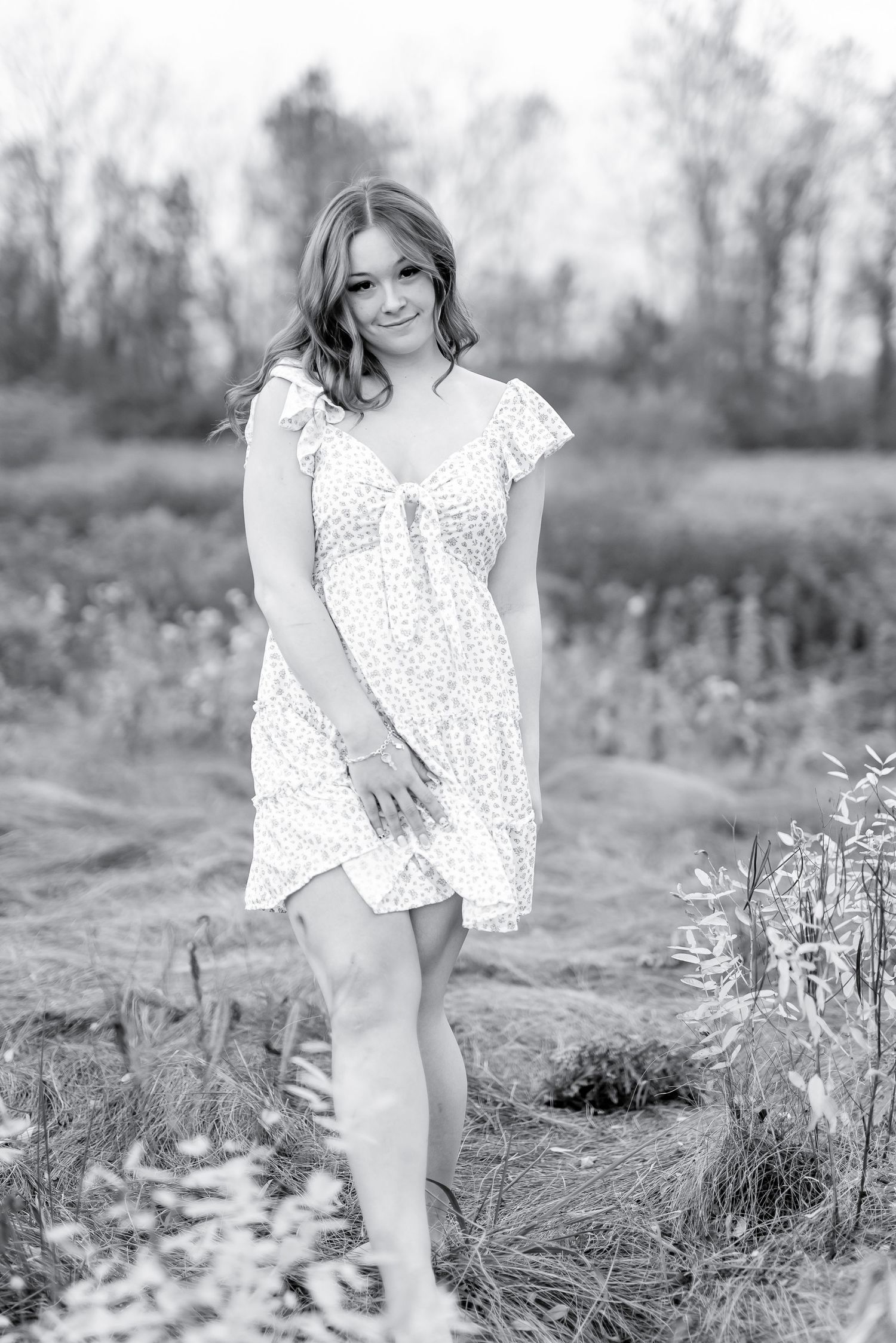 Black and white portrait of smiling young woman in floral sundress standing in a natural field.