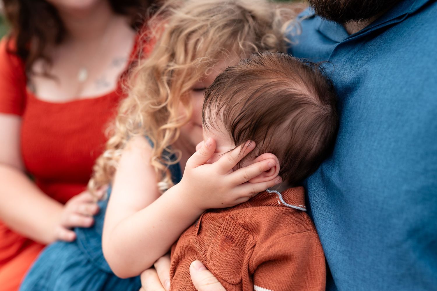 A toddler with curly hair nuzzles a newborn baby held by parents in red and blue clothing.