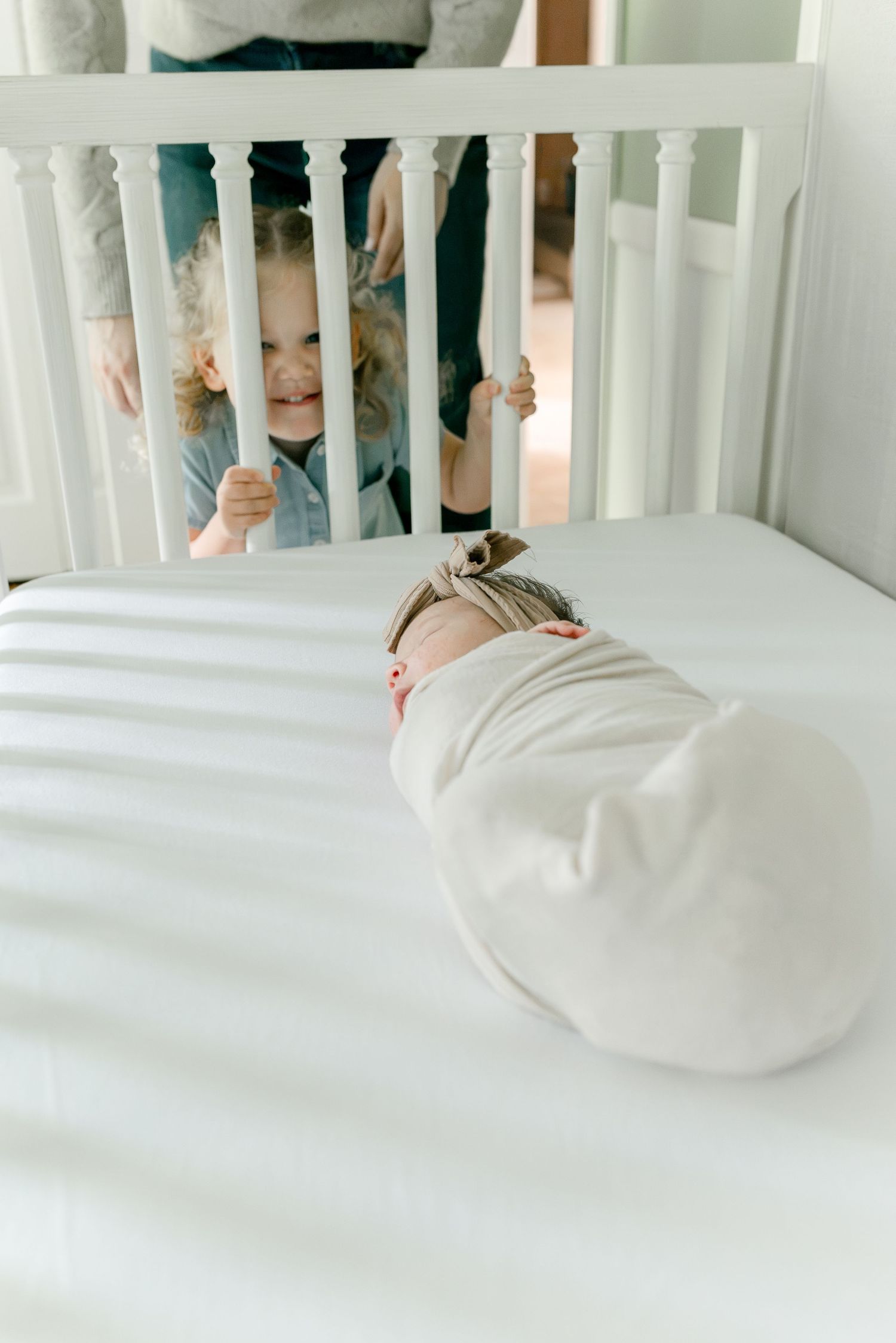 A swaddled newborn lies in a white crib while a smiling toddler peeks through the crib rails.