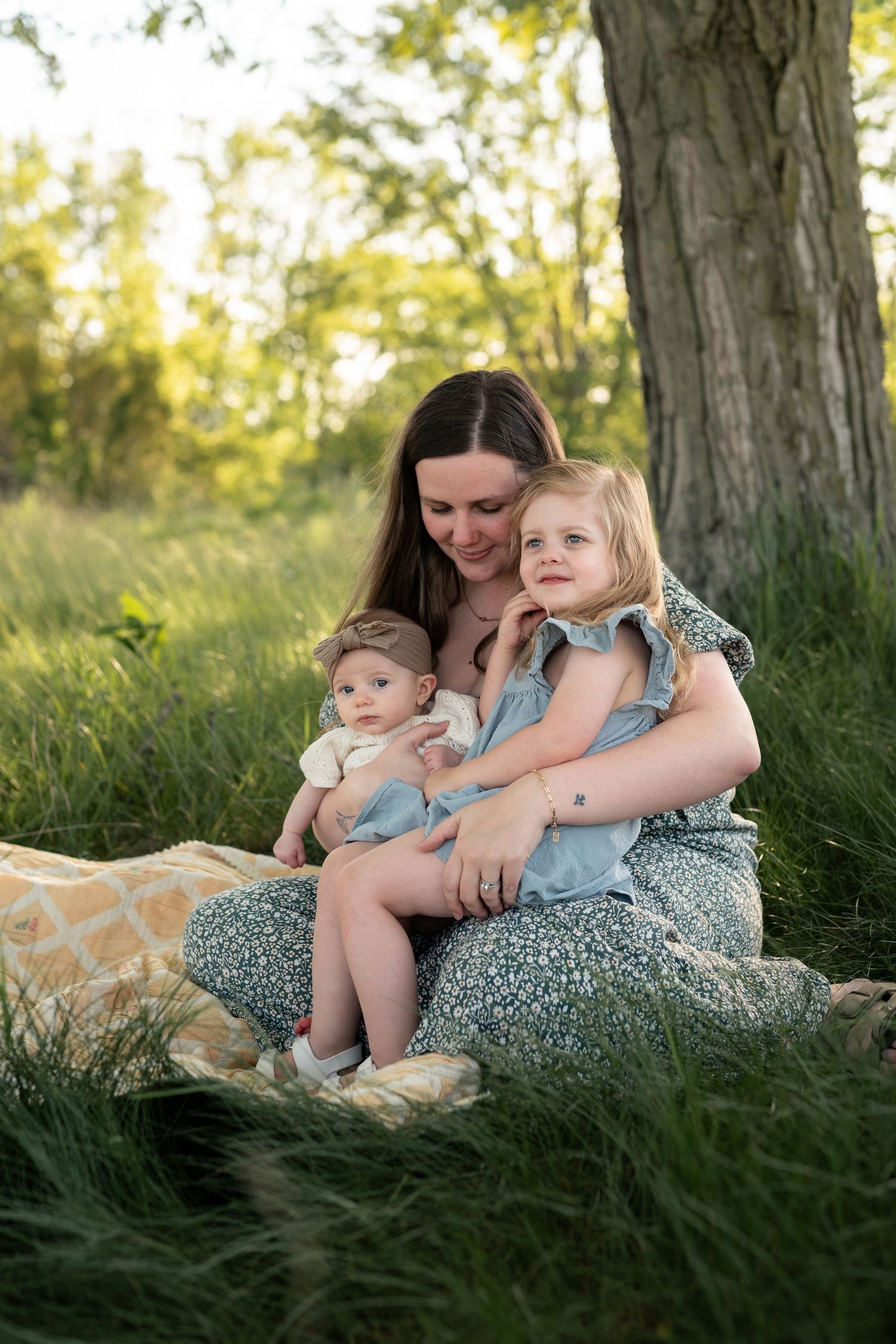 A mother sits on a blanket in green grass, holding her baby and toddler daughter near a tree in golden sunlight.