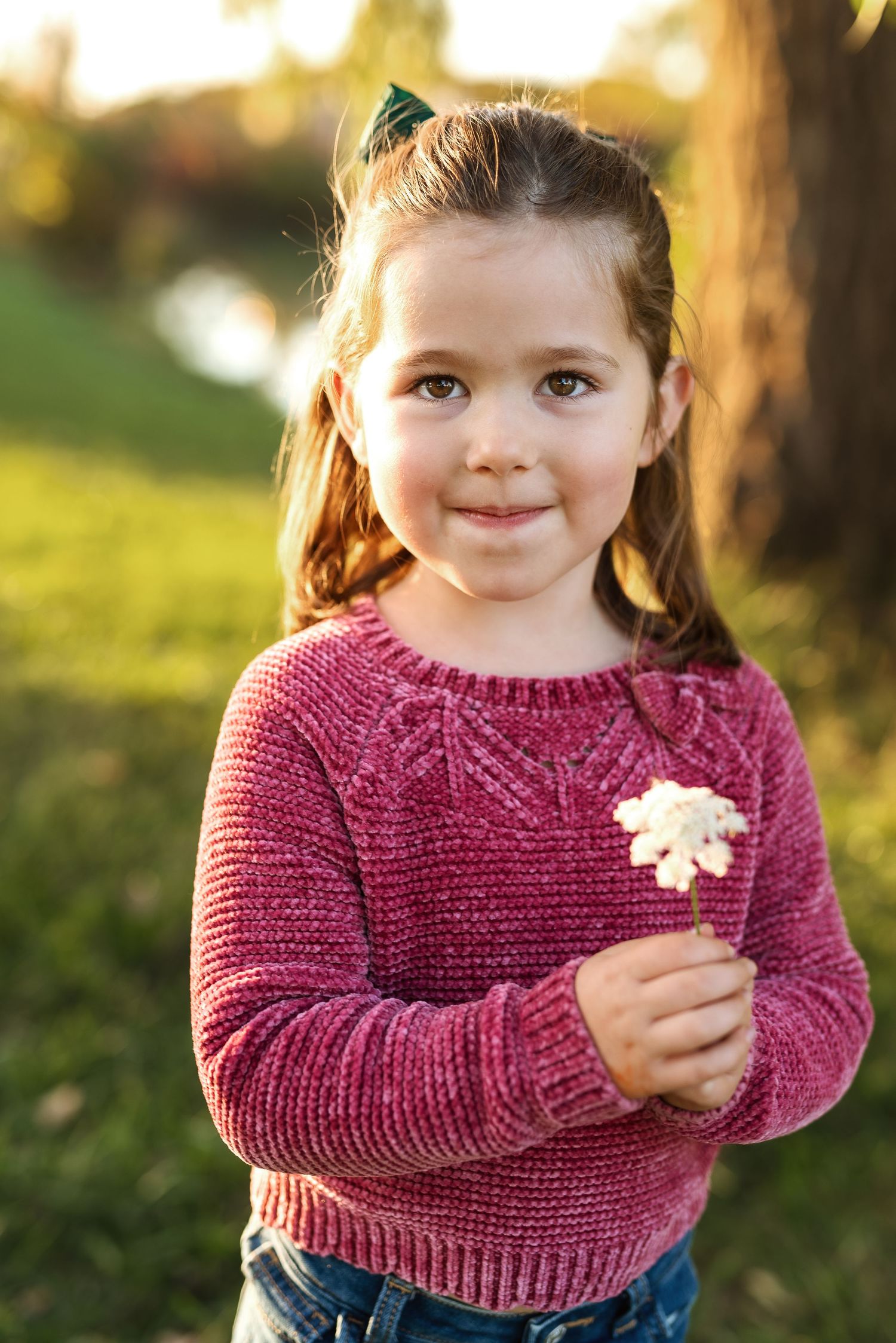 A child in a burgundy sweater holds a leaf while standing outdoors in autumn sunlight.