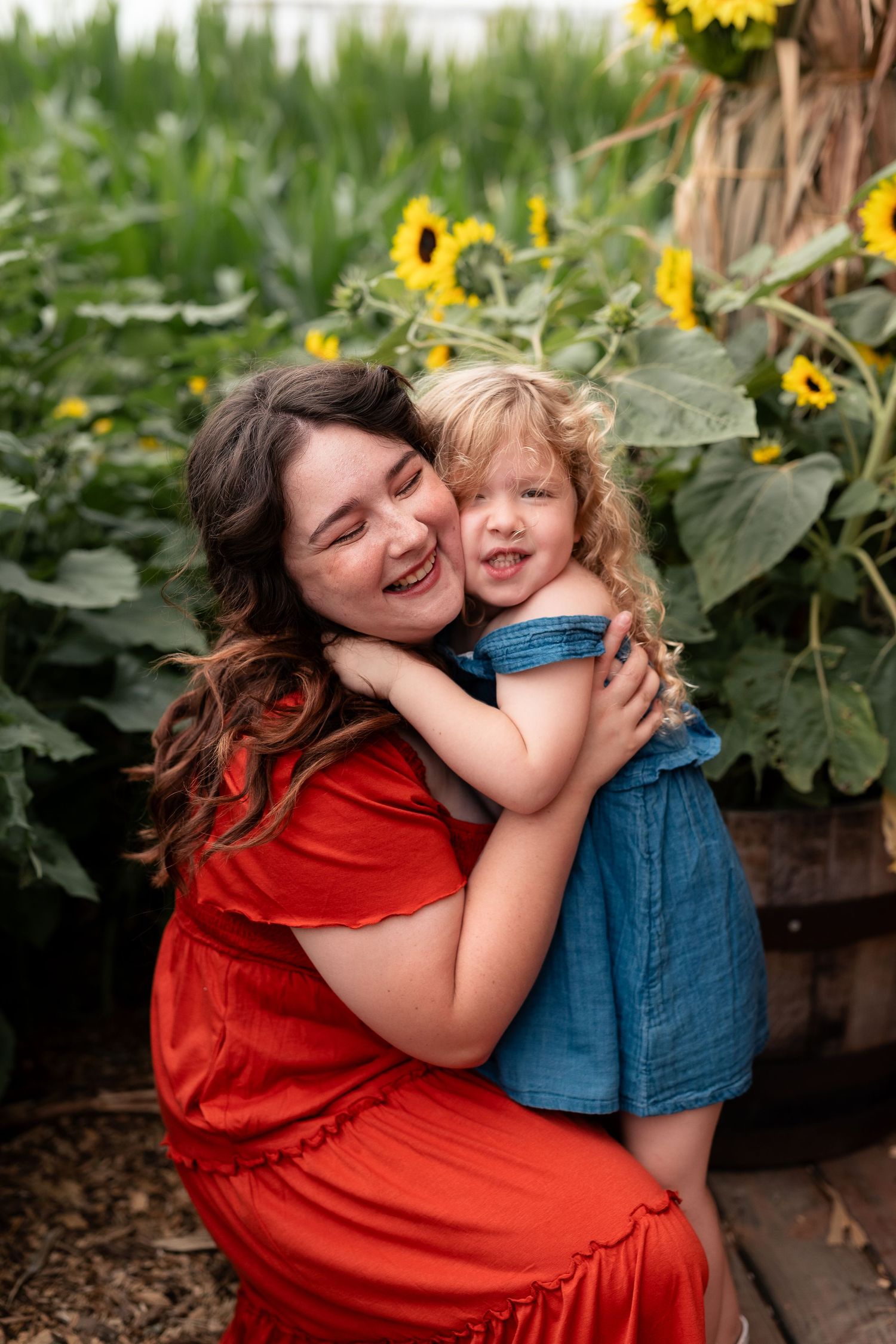 A mother in a red dress hugs her smiling toddler girl in a denim dress among sunflowers.