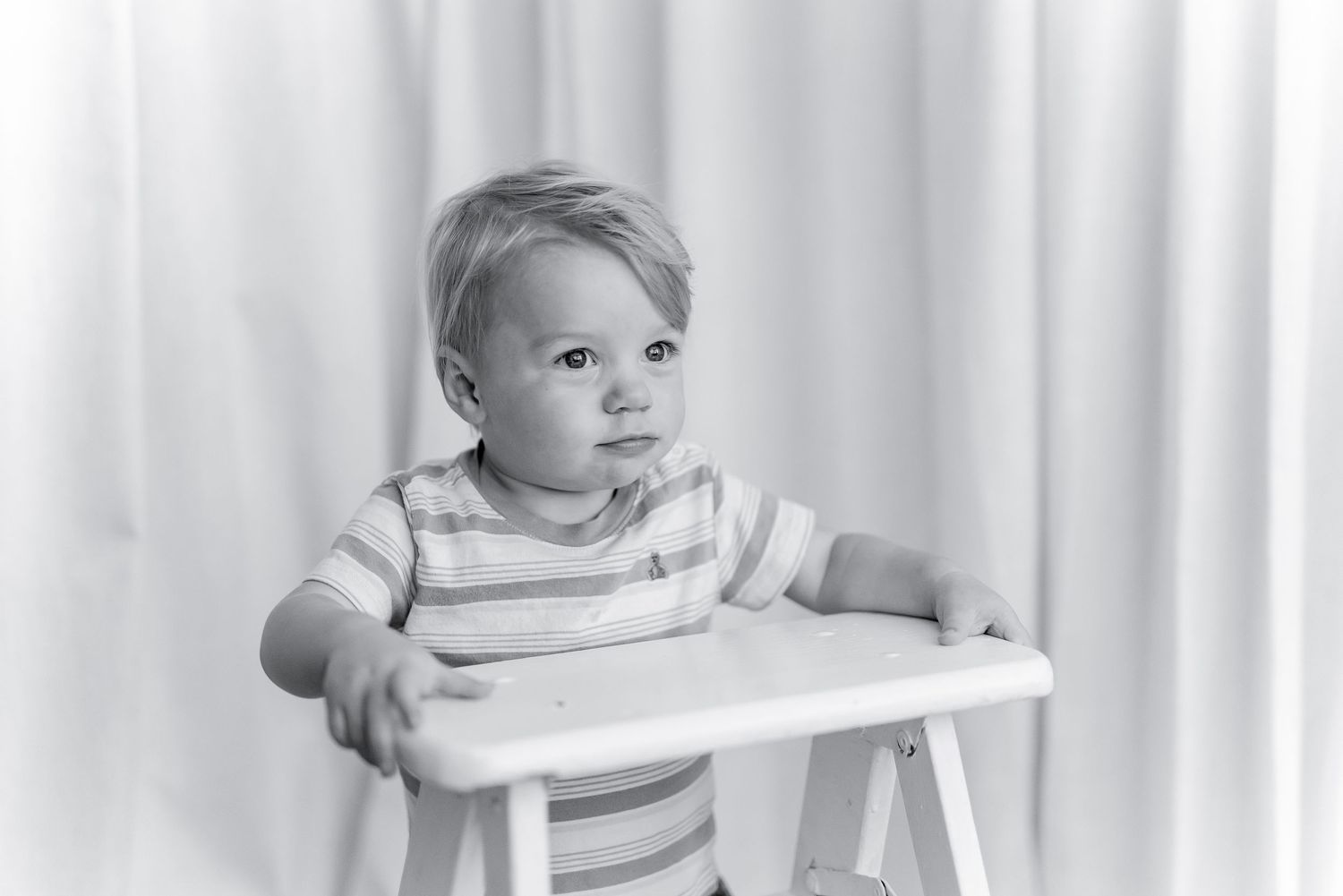 Black and white portrait of a toddler in a striped shirt sitting at a small white wooden table against a soft draped curtain backdrop.