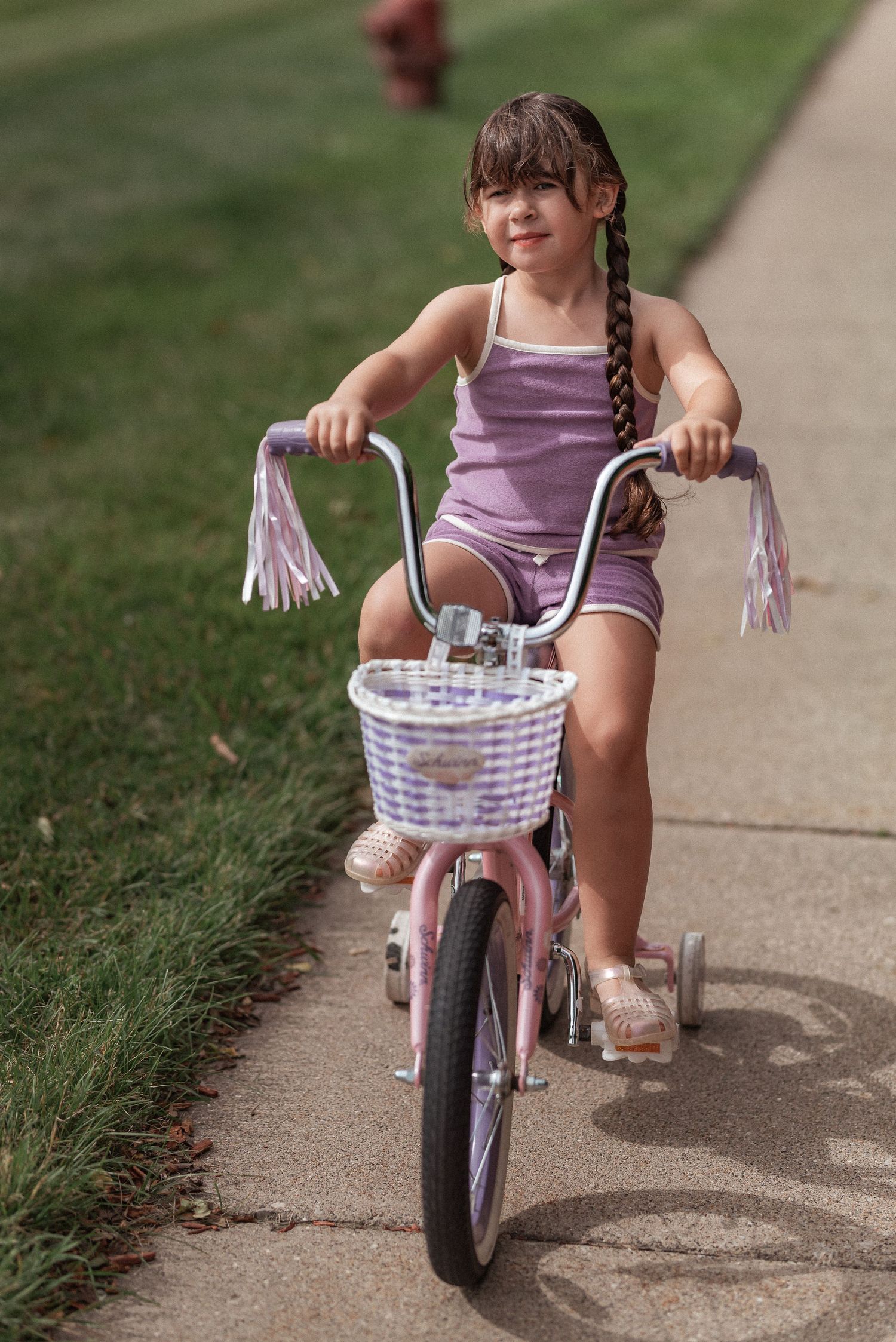 Child on a pink Schwinn bike with streamers and wicker basket riding along a suburban sidewalk.