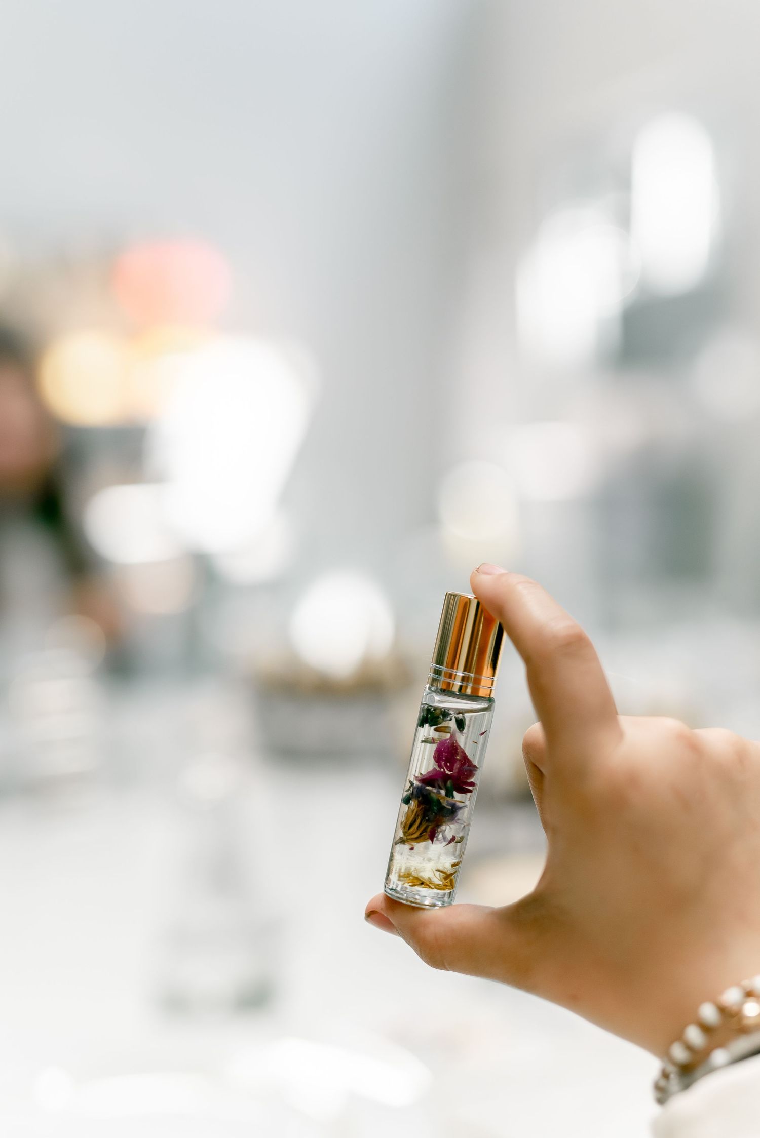 Hand holding a small glass perfume bottle with dried rose petals and gold cap against a blurred bright background.