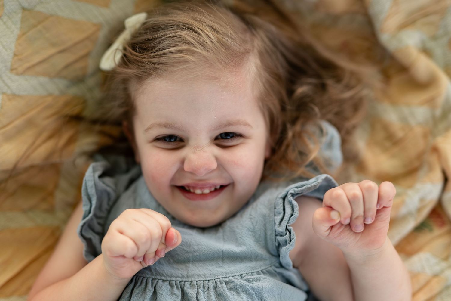 Smiling toddler girl with hair bow lying on a blanket, wearing a gray dress, looking up with a joyful expression.