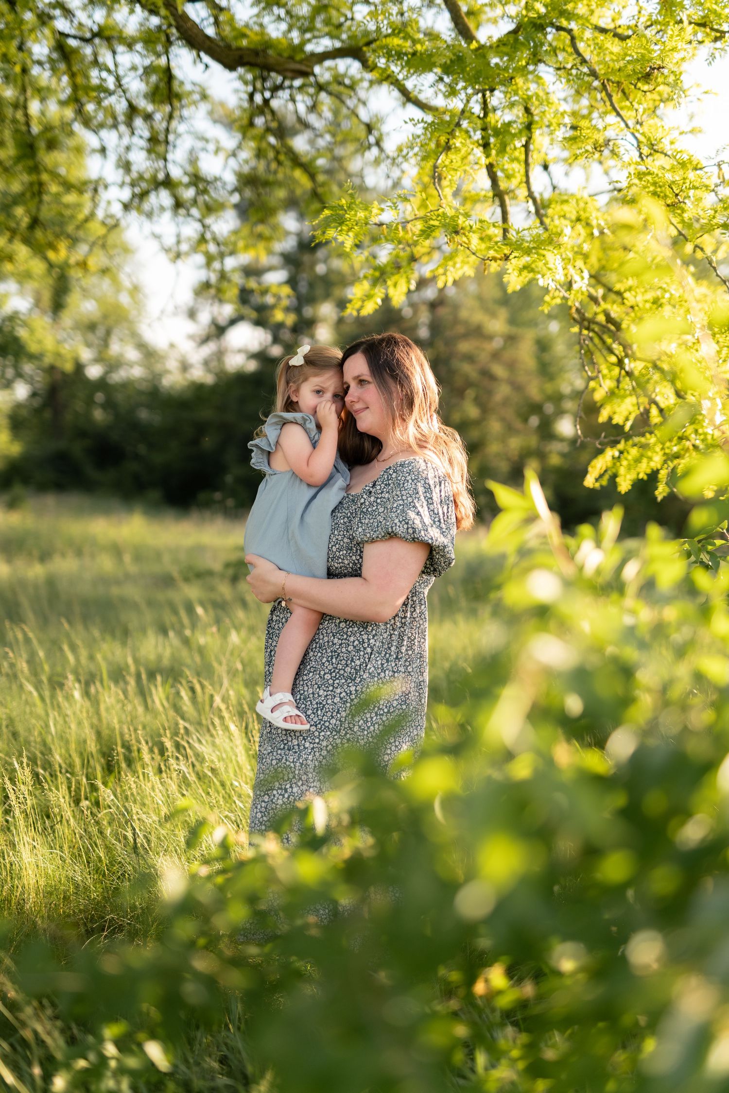A mother holds her toddler in a sunlit green field, both wearing matching gray outfits beneath a leafy tree.