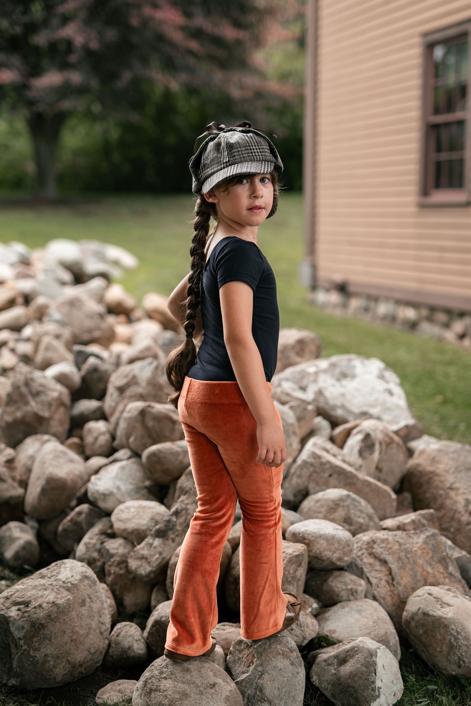 Young girl in orange flare pants, black top, and plaid hat standing on rocks near a yellow house.