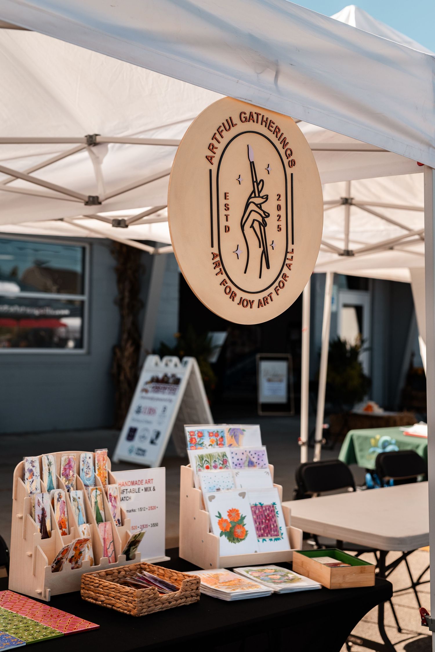 Artful Gatherings art vendor booth displaying colorful cards and prints under a white canopy at an outdoor market.