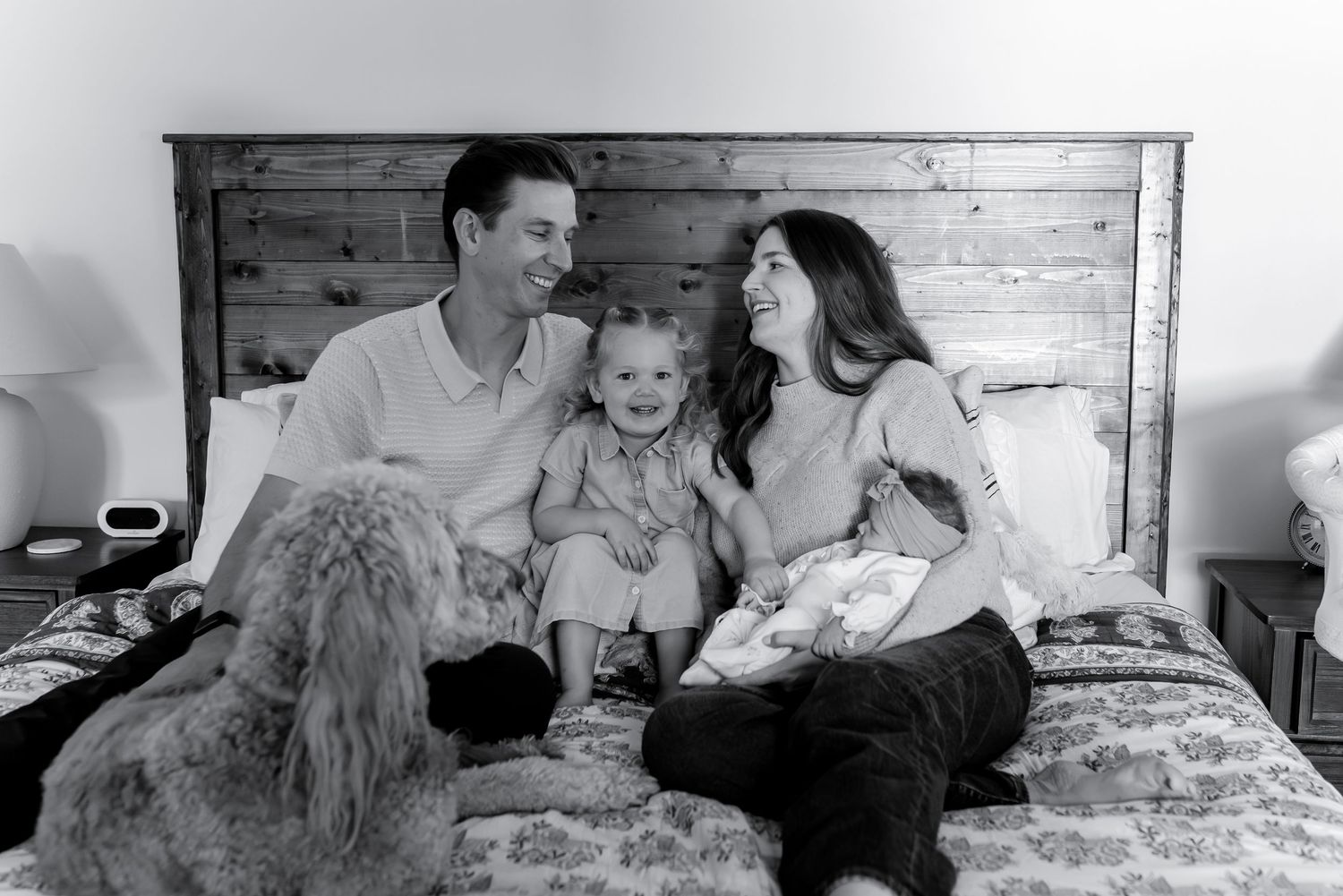 Black and white photo of a happy family with a toddler and fluffy dog relaxing together on a bed.