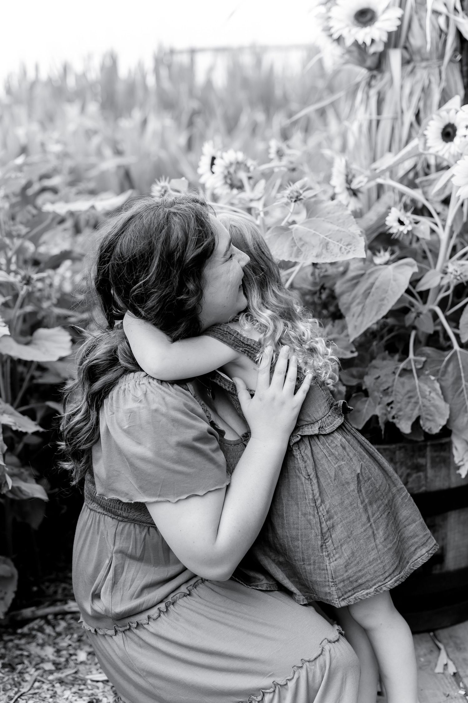 A mother and young daughter share a tender embrace among sunflowers in a black and white garden photo.