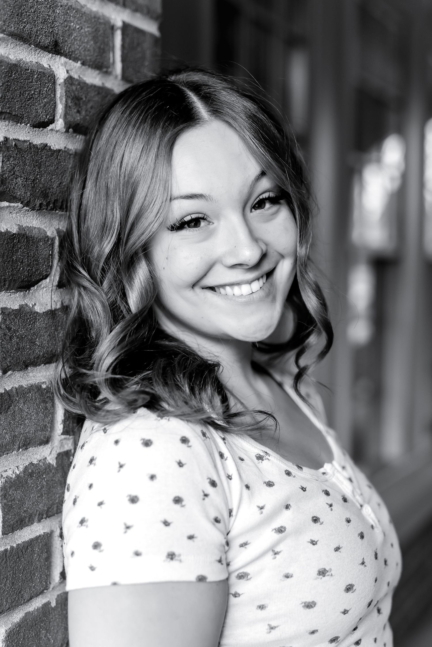 Smiling young woman in polka dot top leaning against brick wall, black and white portrait.