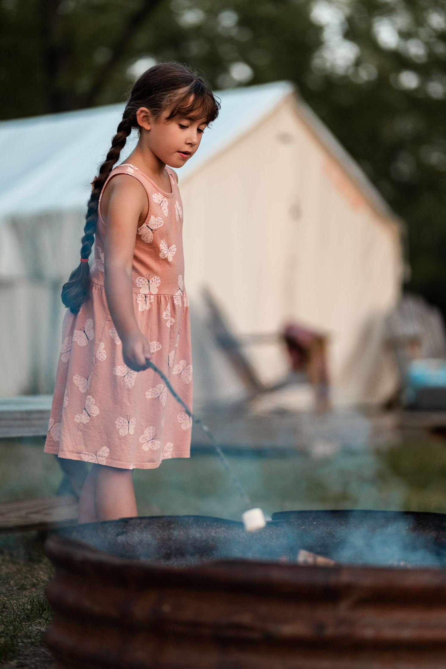 Young girl in pink dress roasting marshmallows over a fire pit at a glamping campsite.