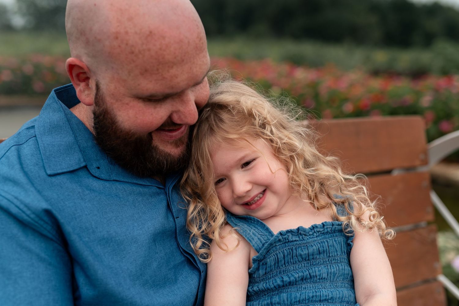 A bald bearded father in blue shirt smiles warmly while holding his curly-haired laughing toddler daughter.