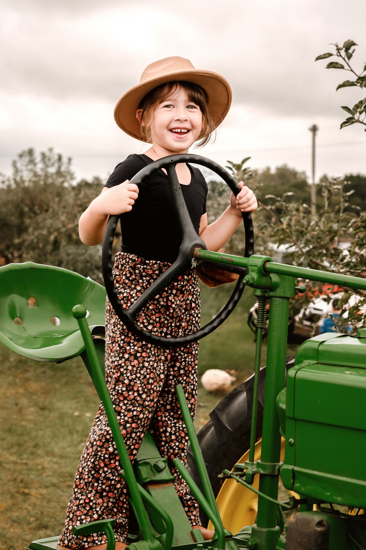 A child in a floral dress and brown hat sits playfully on a vintage green John Deere tractor in a rustic outdoor setting.