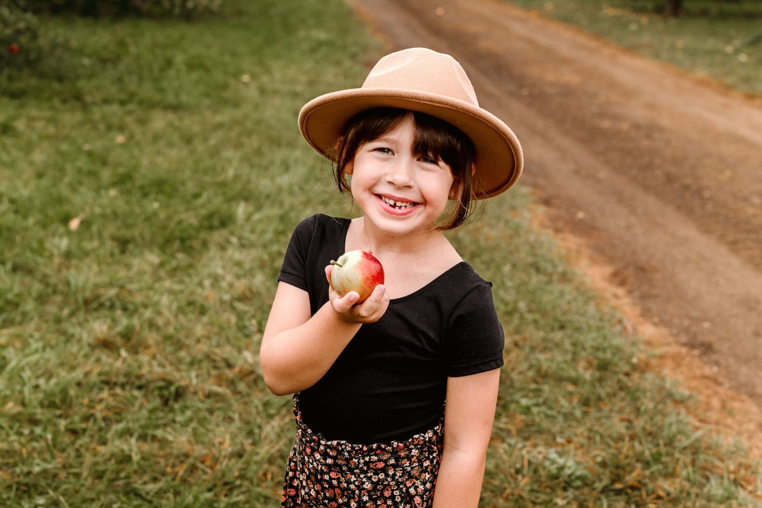 A person wearing a brown hat and black shirt poses playfully on a dirt path surrounded by green grass.