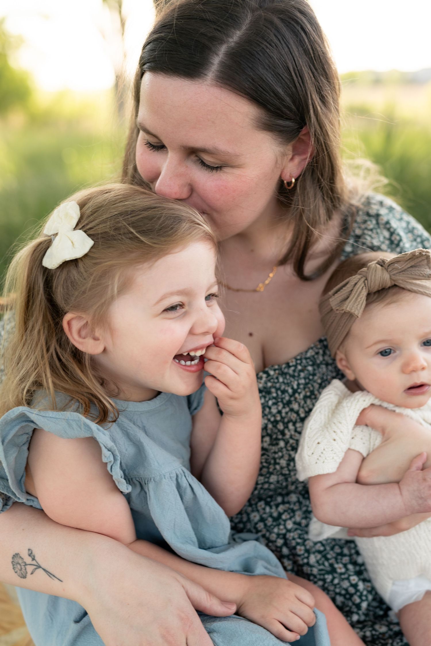 A mother smiles warmly while holding her laughing toddler daughter and infant baby outdoors in a sunlit field.