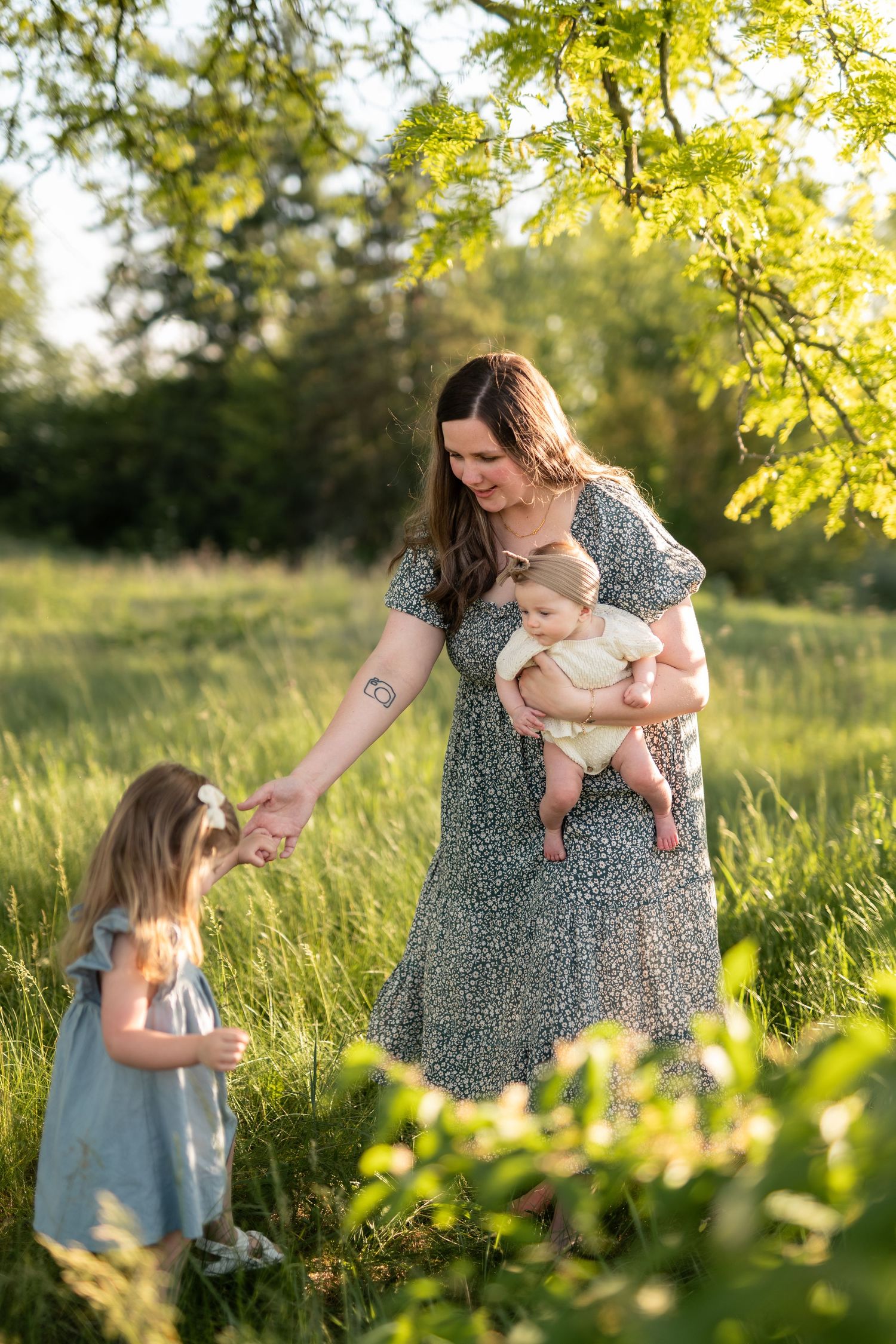 A mother in a floral dress holds her baby while holding hands with her toddler daughter in a sunny green field.