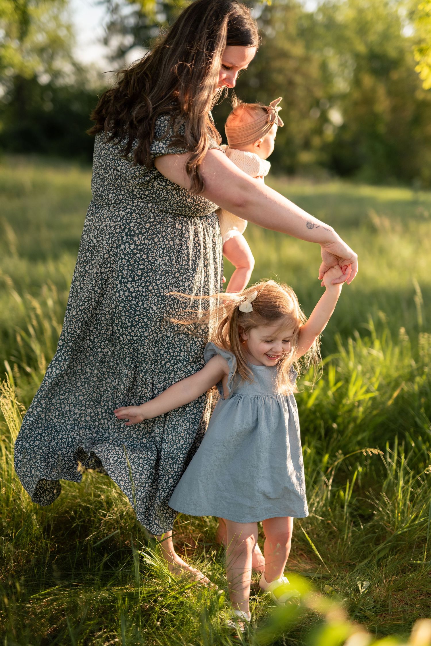 Mother in floral dress dancing with two young daughters in sunlit green field during golden hour.
