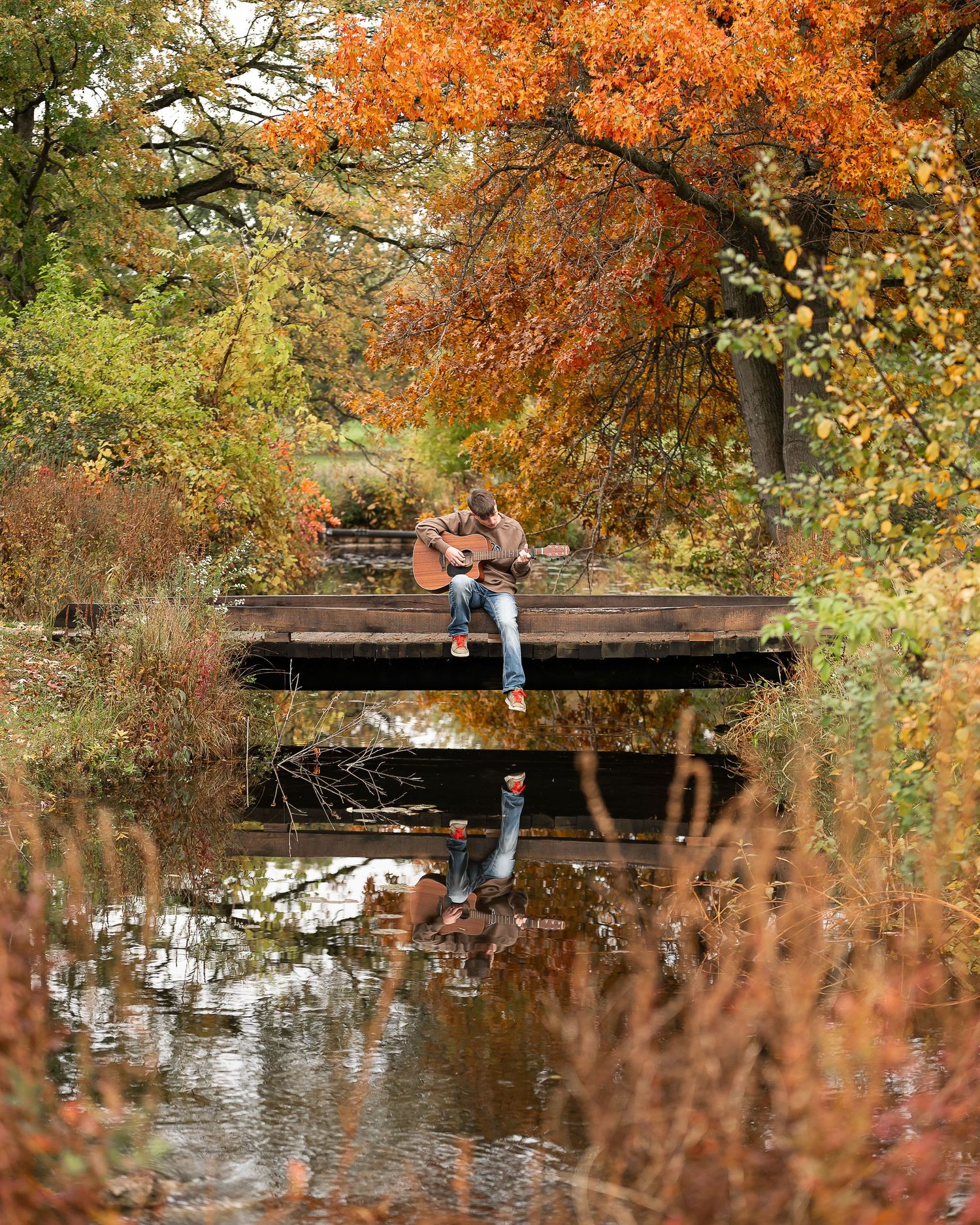 Person playing guitar on a wooden bridge surrounded by vibrant autumn foliage reflecting in calm water below.