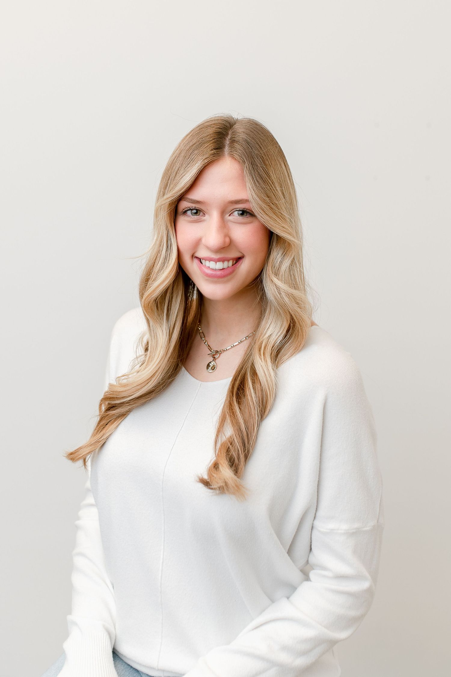 Young blonde woman smiling in white long-sleeve top with necklace against light gray background.
