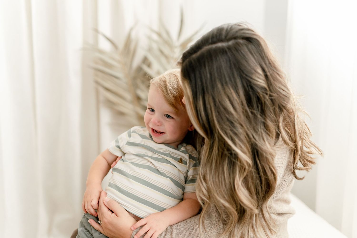 A mother with long wavy hair lovingly holds her smiling toddler in a bright, airy white room.
