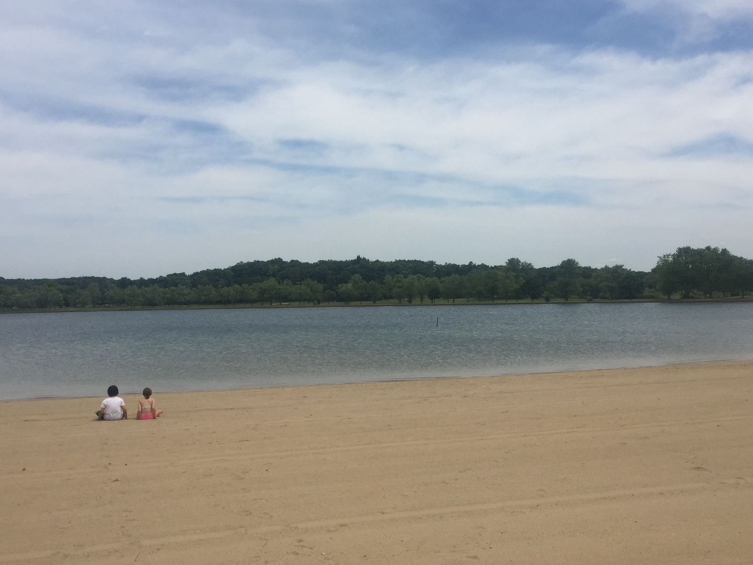 Two people sit on a sandy beach beside a calm lake surrounded by lush green trees under a partly cloudy sky.