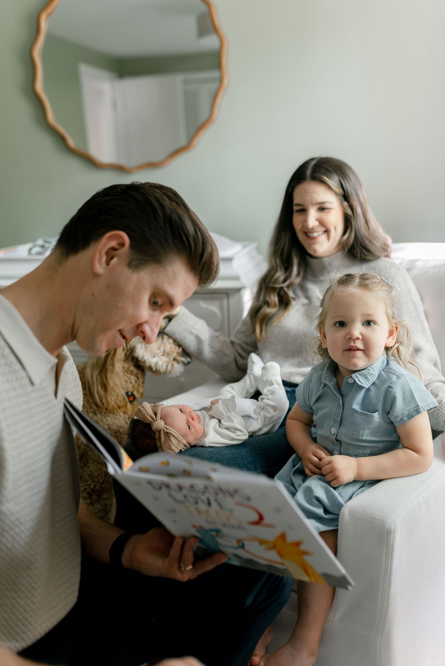 Family of four sits on couch as dad reads a picture book to newborn baby and toddler daughter.