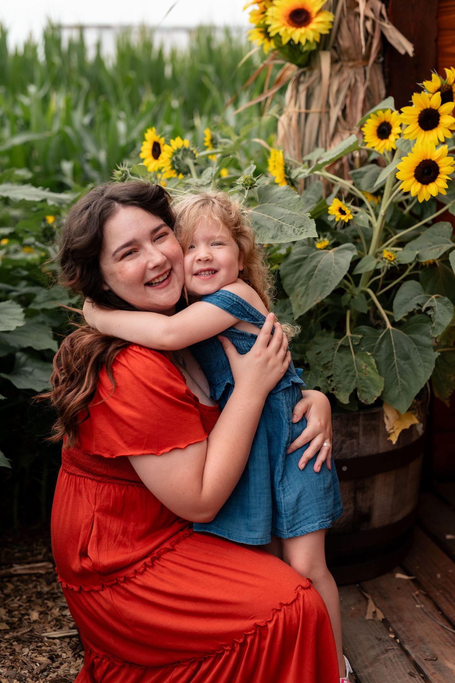 A laughing woman in a red dress hugs a smiling toddler girl near sunflowers in a garden setting.
