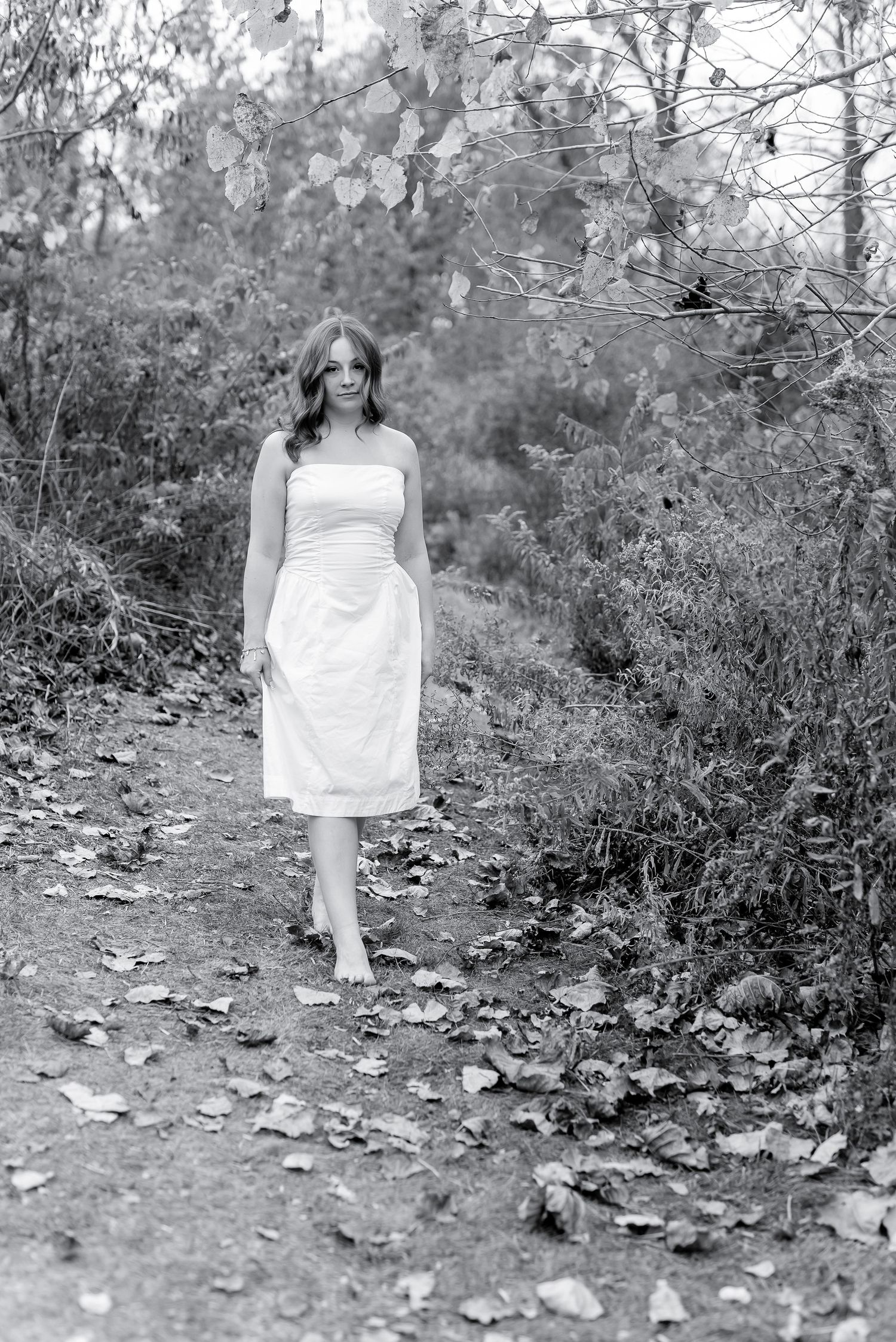 Black and white photo of barefoot woman in white dress walking on leaf-covered forest path.