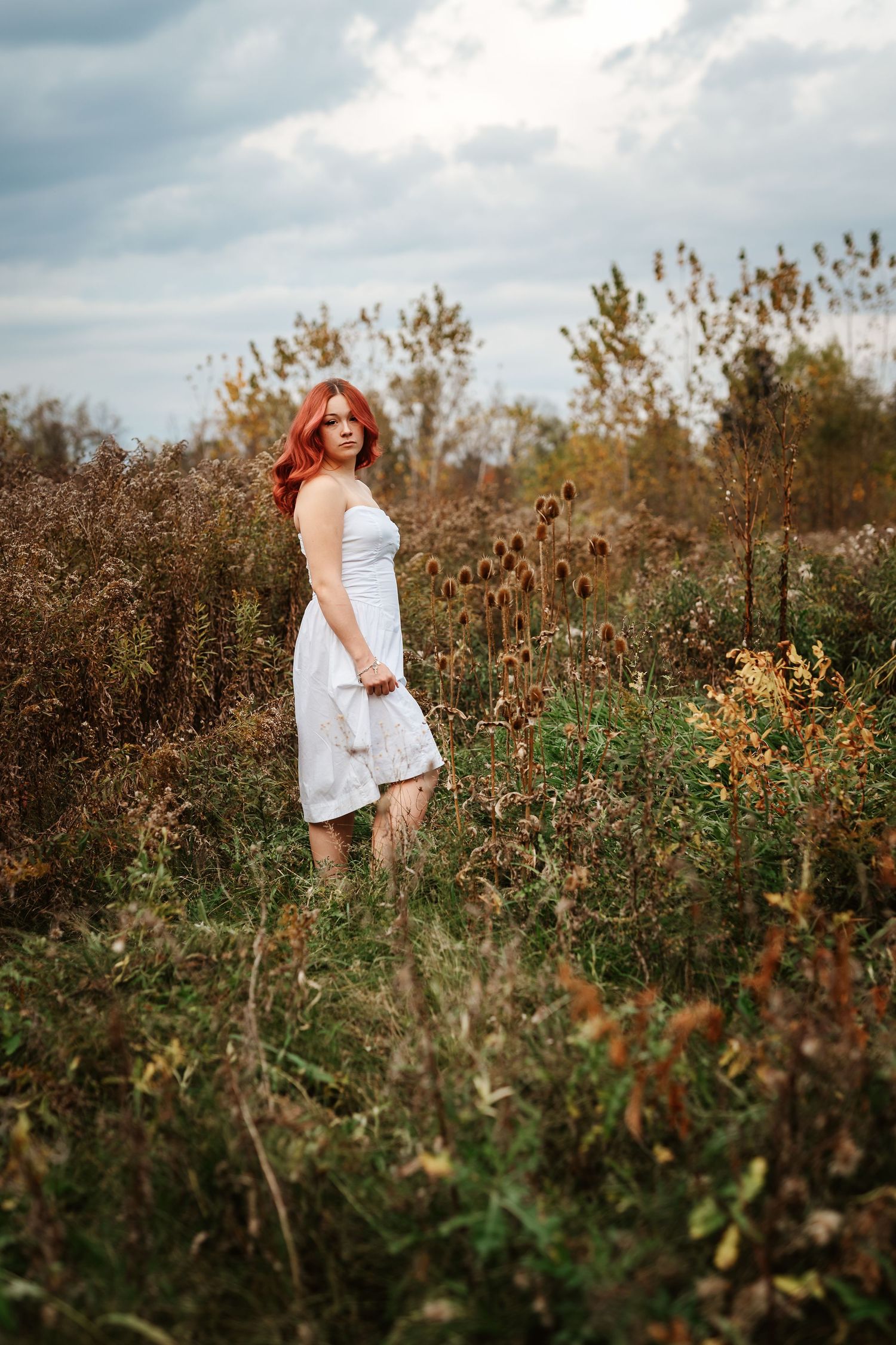 Red-haired woman in white dress standing in autumn field with dried wildflowers under cloudy sky.