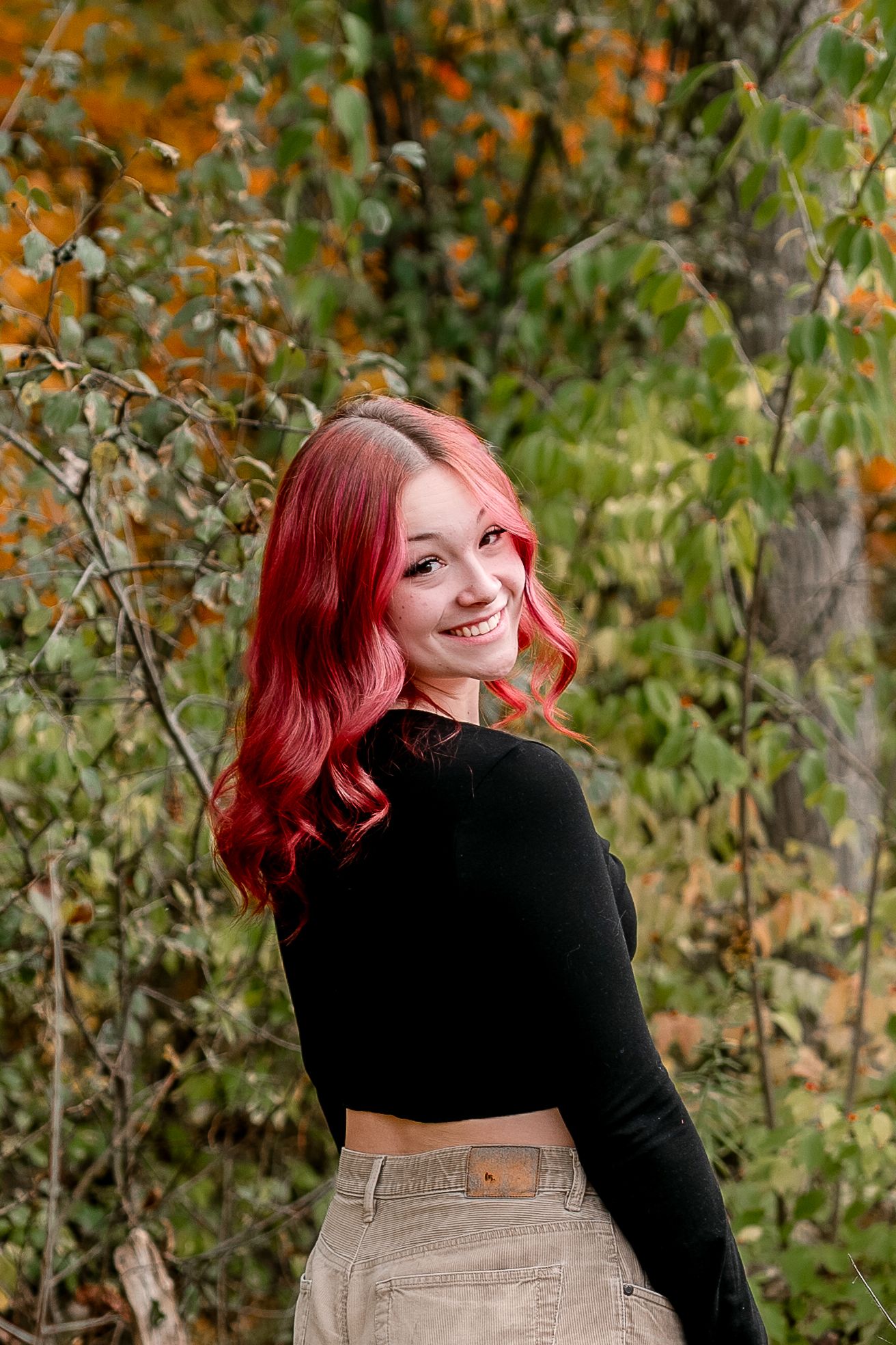 Young woman with vibrant red hair smiles over shoulder wearing black crop top amid colorful autumn foliage.