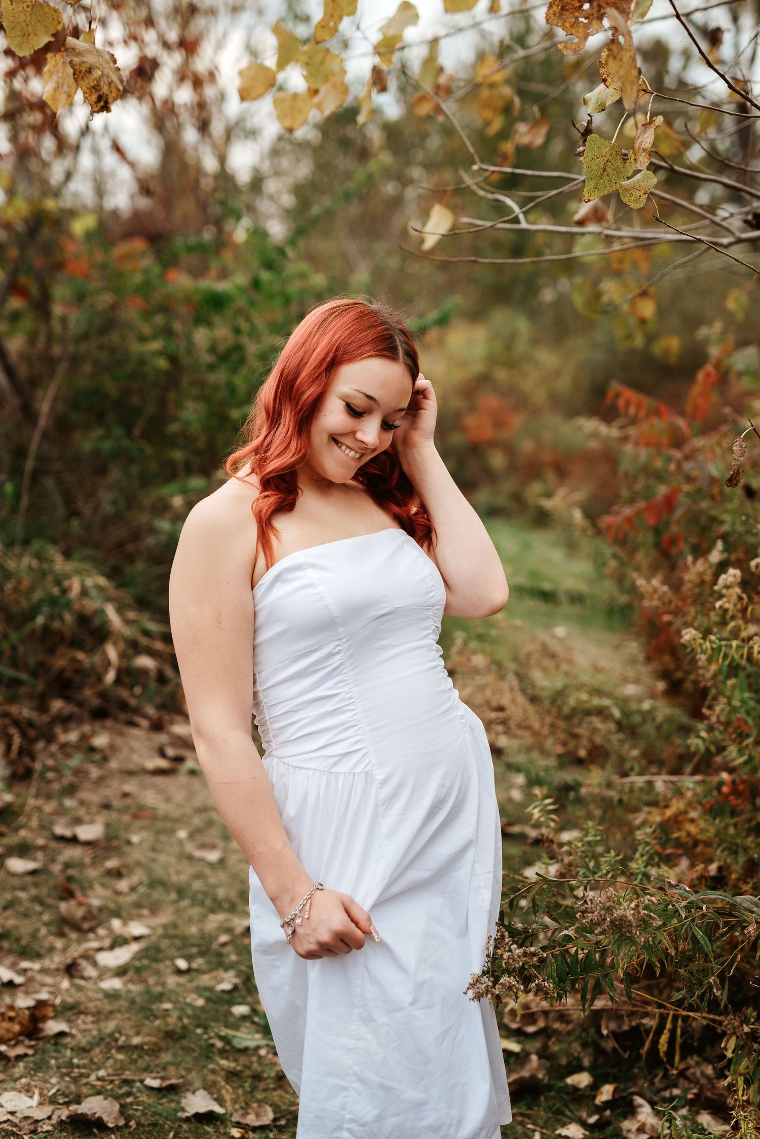 Smiling redhead woman in white strapless dress poses outdoors among autumn foliage and fallen leaves.