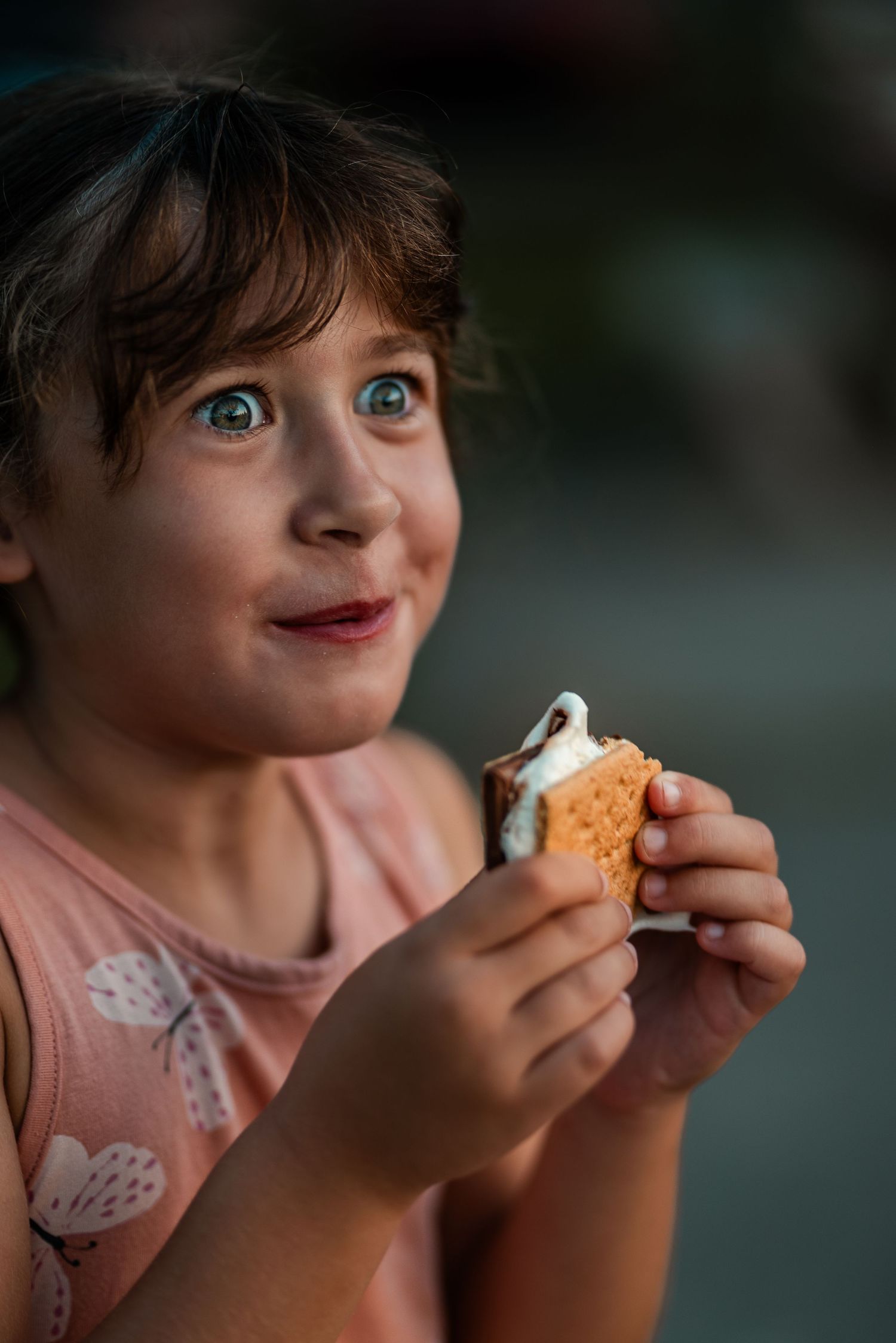 Young girl with wide eyes holding a s'more treat, wearing a pink shirt, looking excited while eating outdoors.