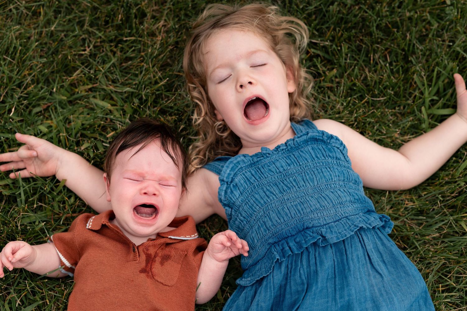 Two young siblings lying on green grass, both laughing with arms outstretched in pure joy.