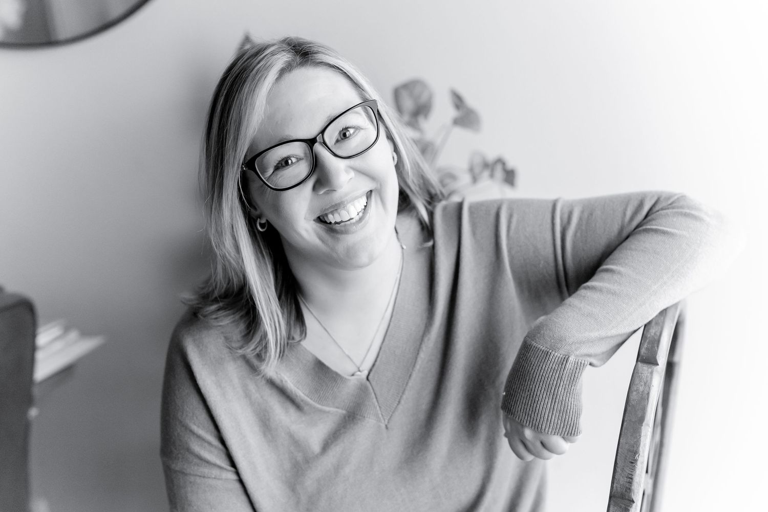 Black and white portrait of someone wearing glasses and a sweater leaning on a chair while smiling warmly.