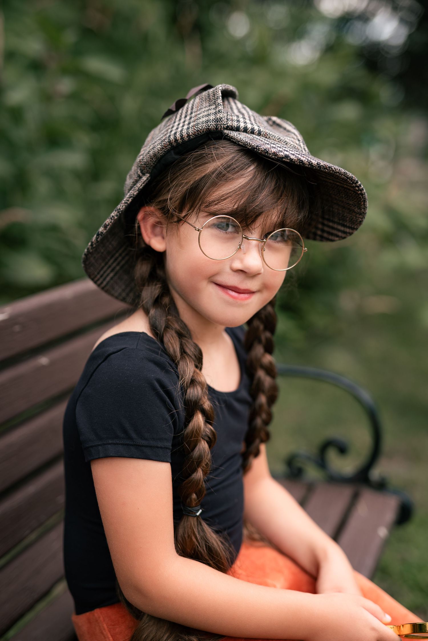Young girl with braids, round glasses, and plaid cap sits smiling on a park bench in a black shirt.