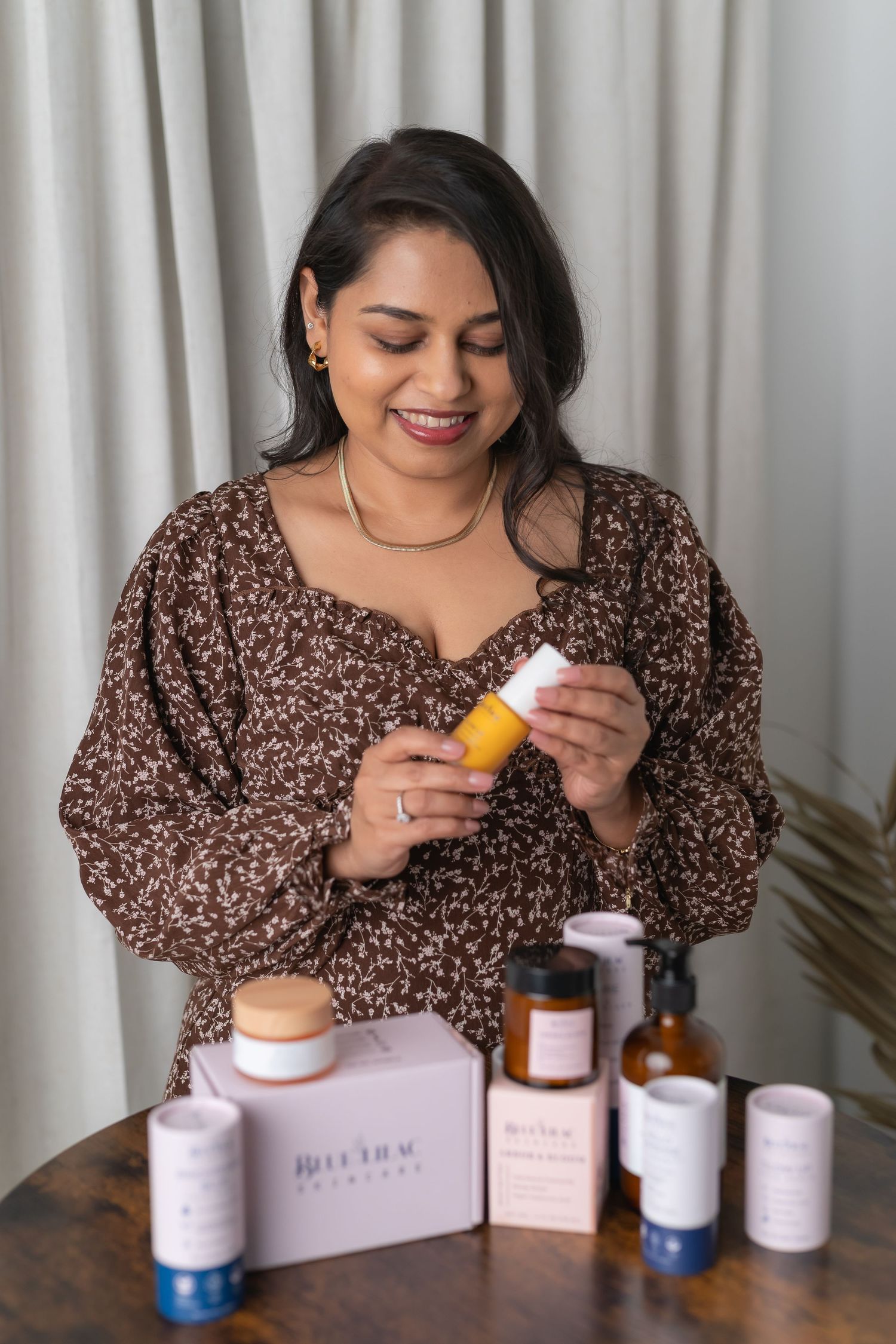 Smiling woman in floral dress examining skincare products from a Blue Hue Naturals beauty box on a wooden table.