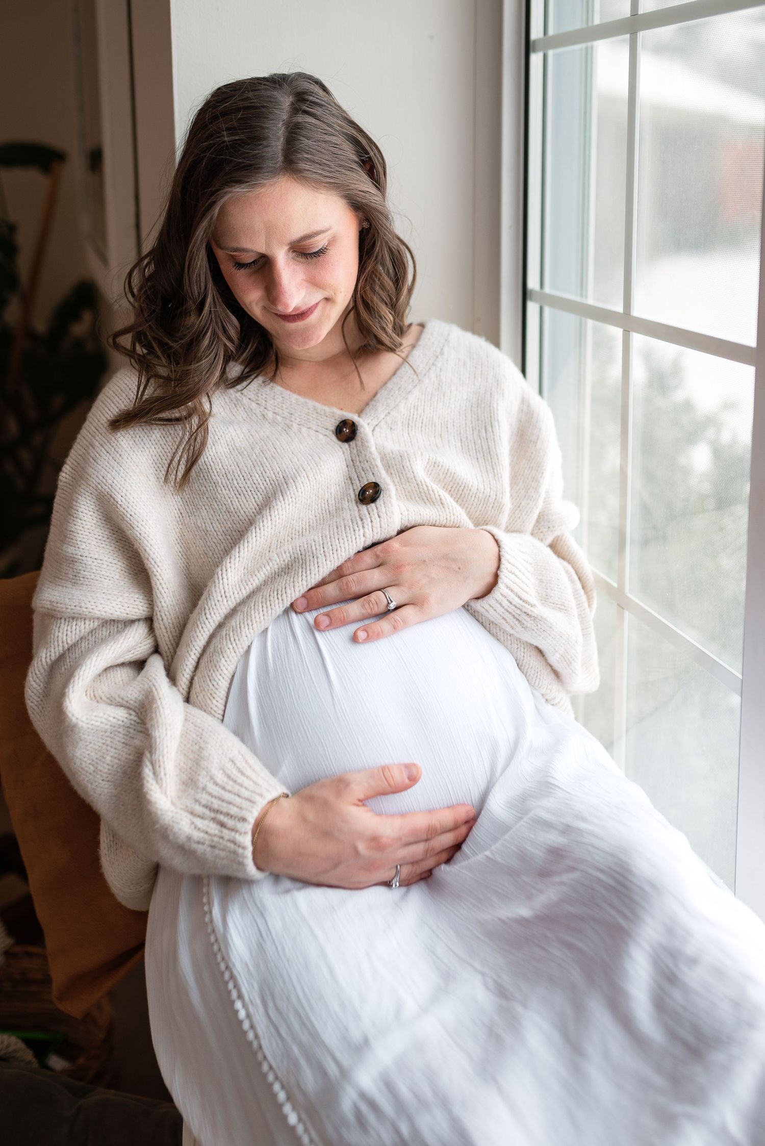 Serene maternity moment by window in cozy white cardigan.