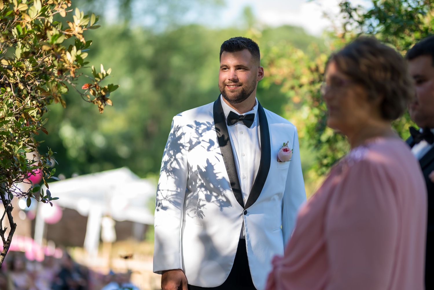 An elegant groom stands at a wedding ceremony wearing a crisp white tuxedo jacket and black bow tie in a sunny garden setting.