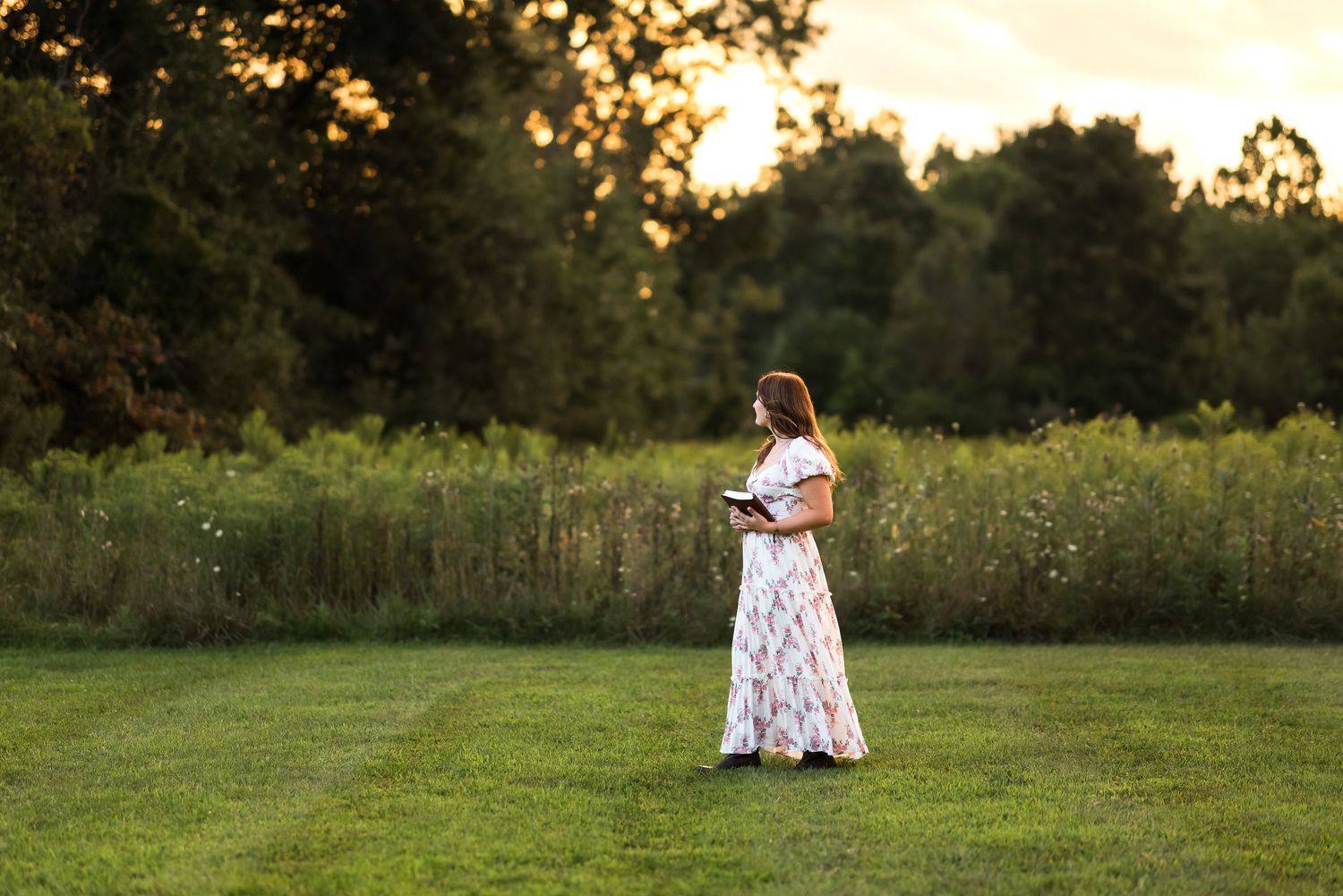 A person in a white dress stands in a grassy field at golden hour during sunset.