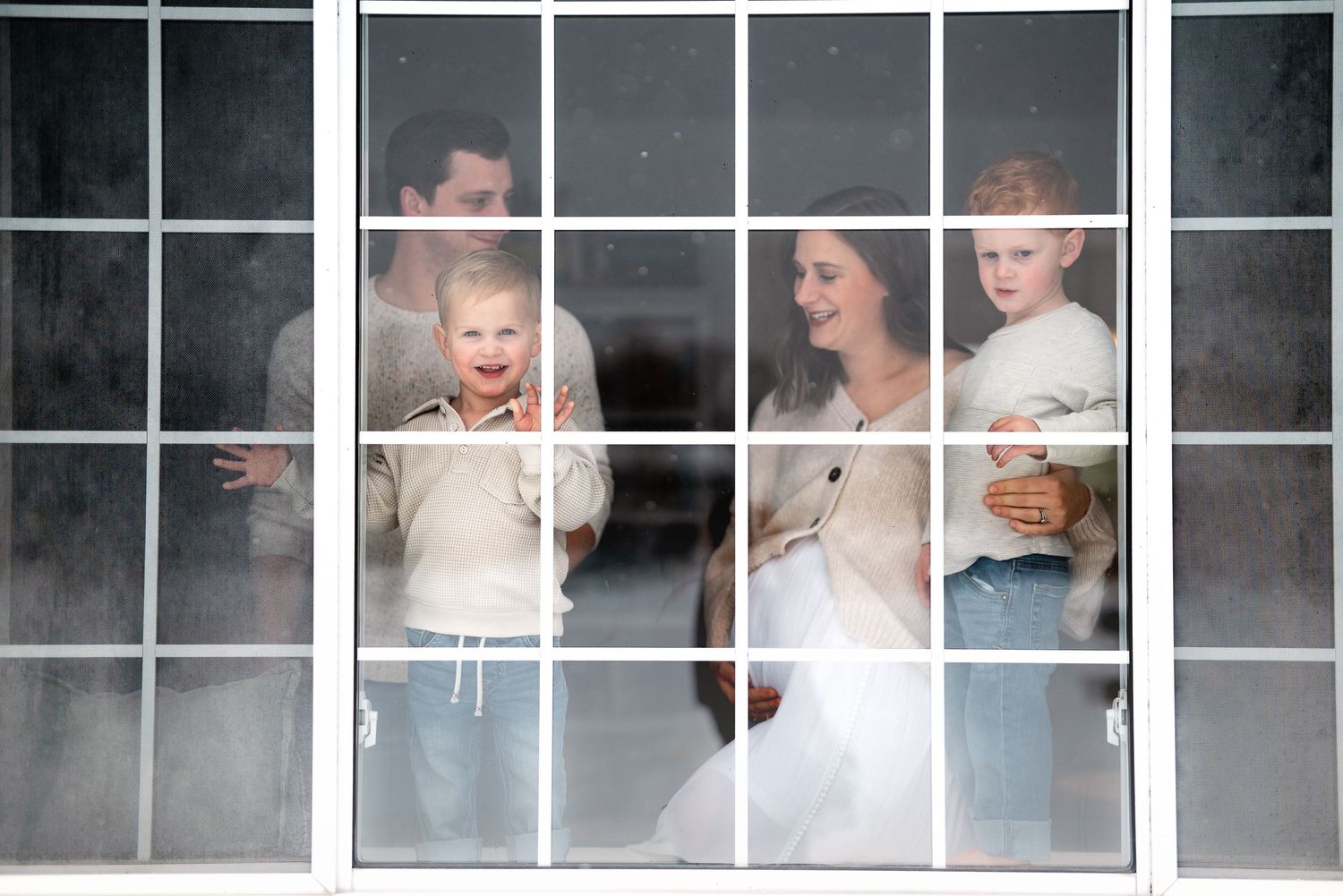Family of four posing behind a white-framed window, parents holding young children in a candid portrait photo.