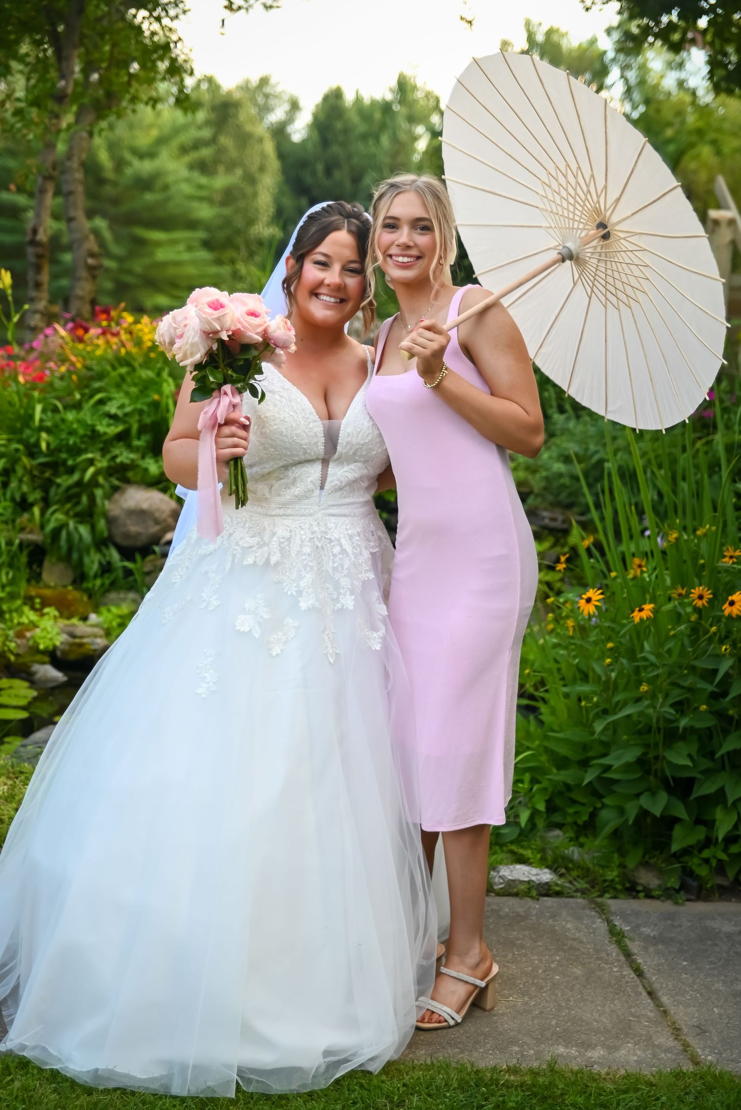 A bride in a white ballgown and her companion in a pink dress pose with a decorative parasol in a garden setting.