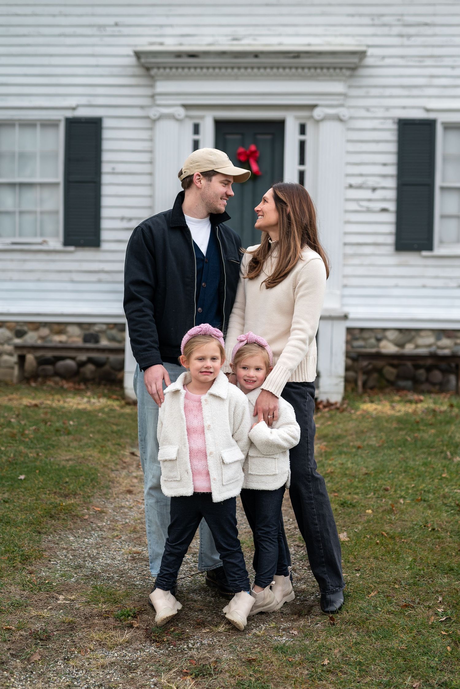 A family portrait capturing a casual moment outside a historic white farmhouse with black shutters.