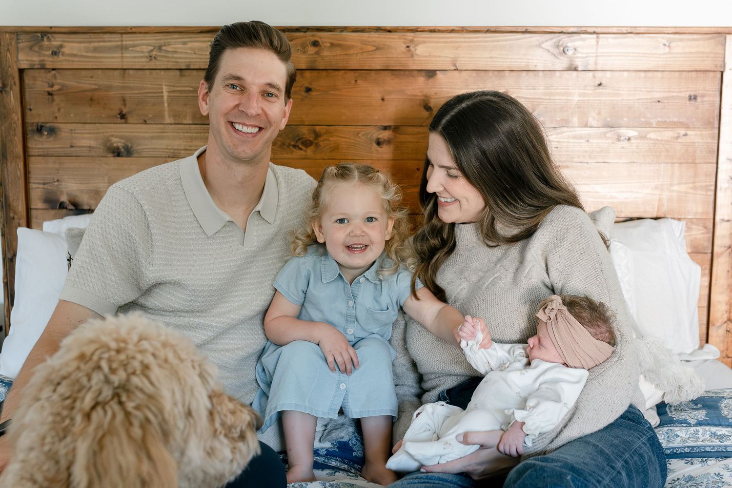 Happy family of four with a toddler and newborn baby sitting on a bed with a golden dog.