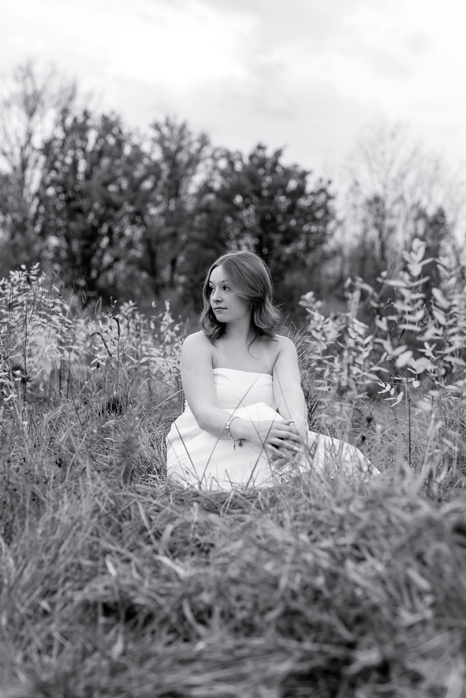 Black and white image of young woman in white dress sitting peacefully among tall meadow grasses.