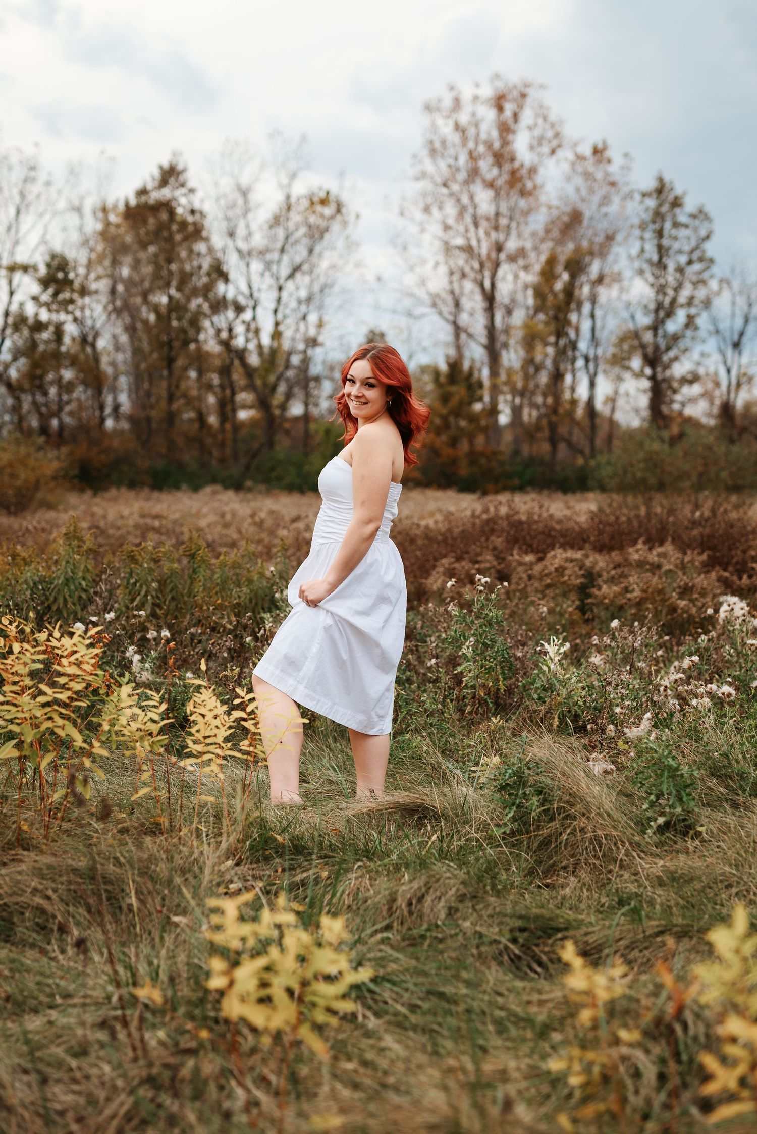 Red-haired woman in white dress standing in autumn field with wildflowers and trees.
