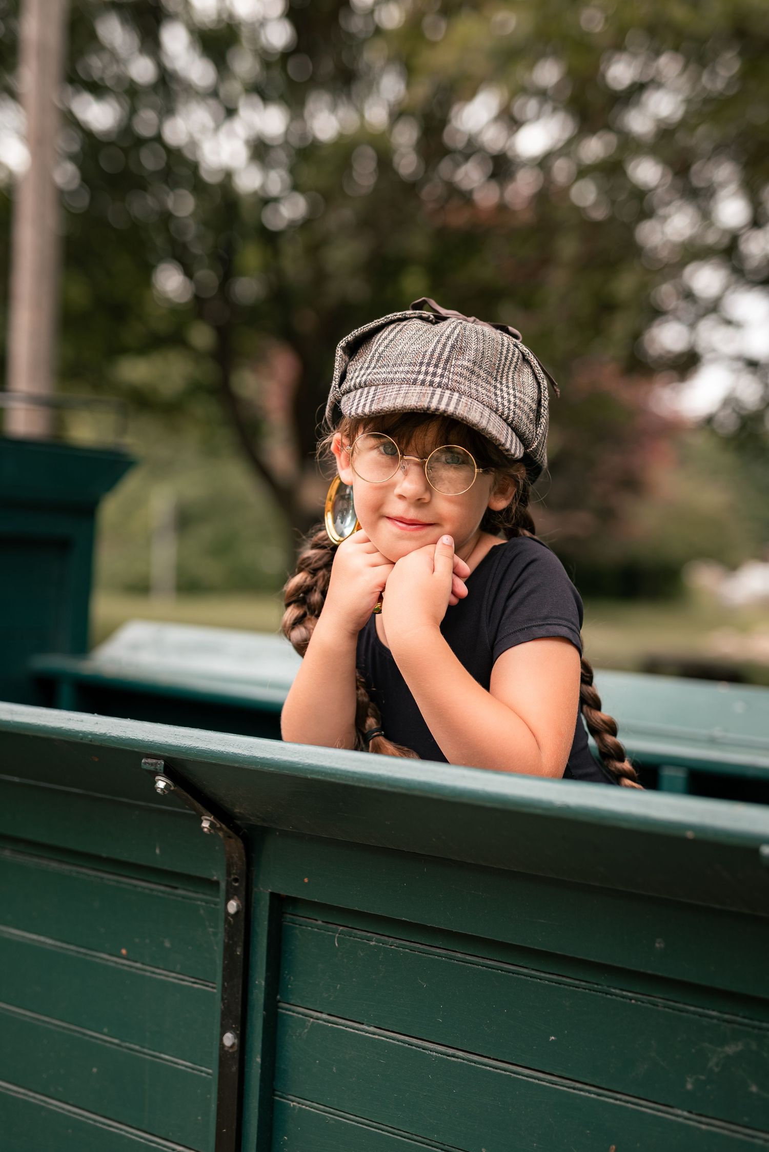 Young girl with braids, plaid cap, and glasses resting chin on hands atop a green wooden structure outdoors.