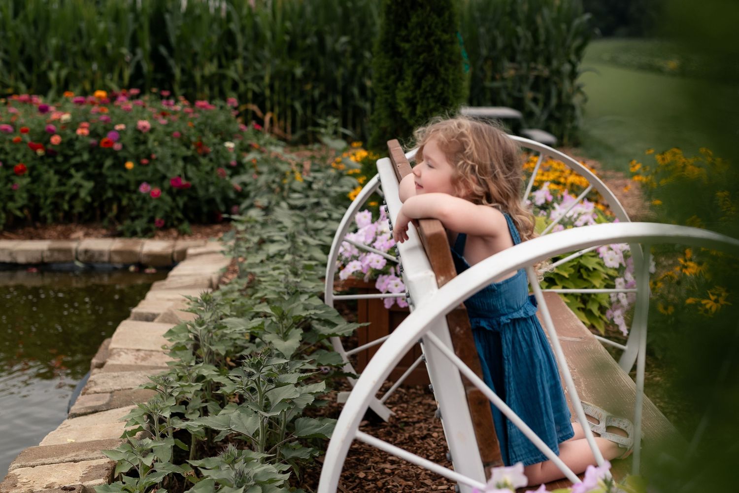 Young girl in blue dress leans over white garden bridge surrounded by colorful flowers and lush greenery.